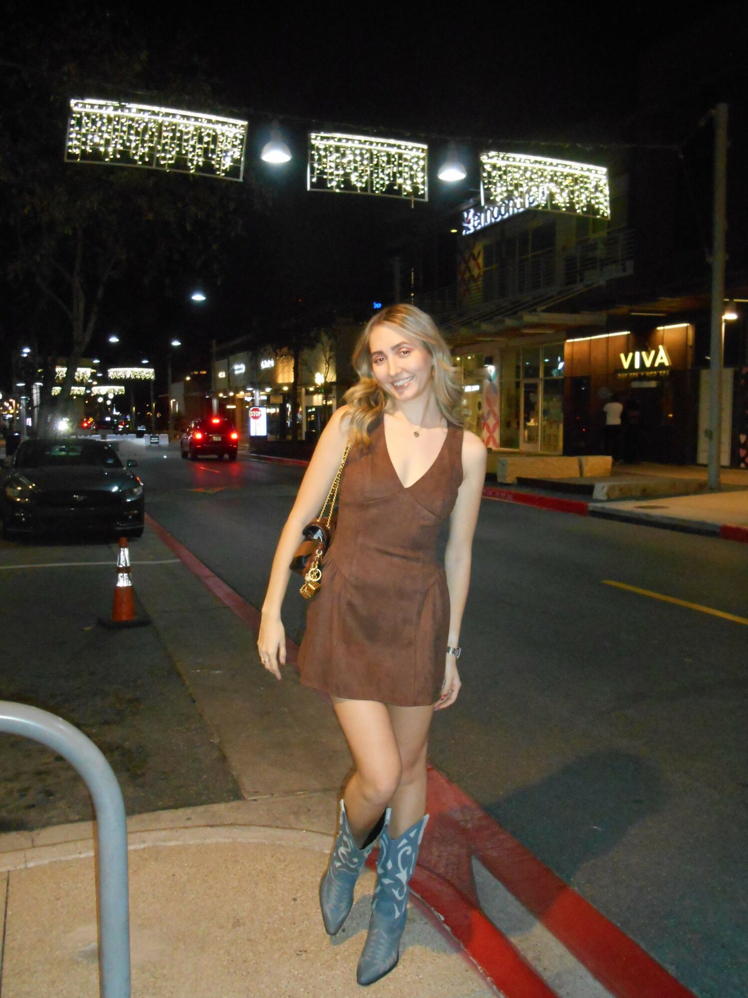 Young woman standing on sidewalk at night, wearing a brown dress and blue shoes, illuminated by streetlights.