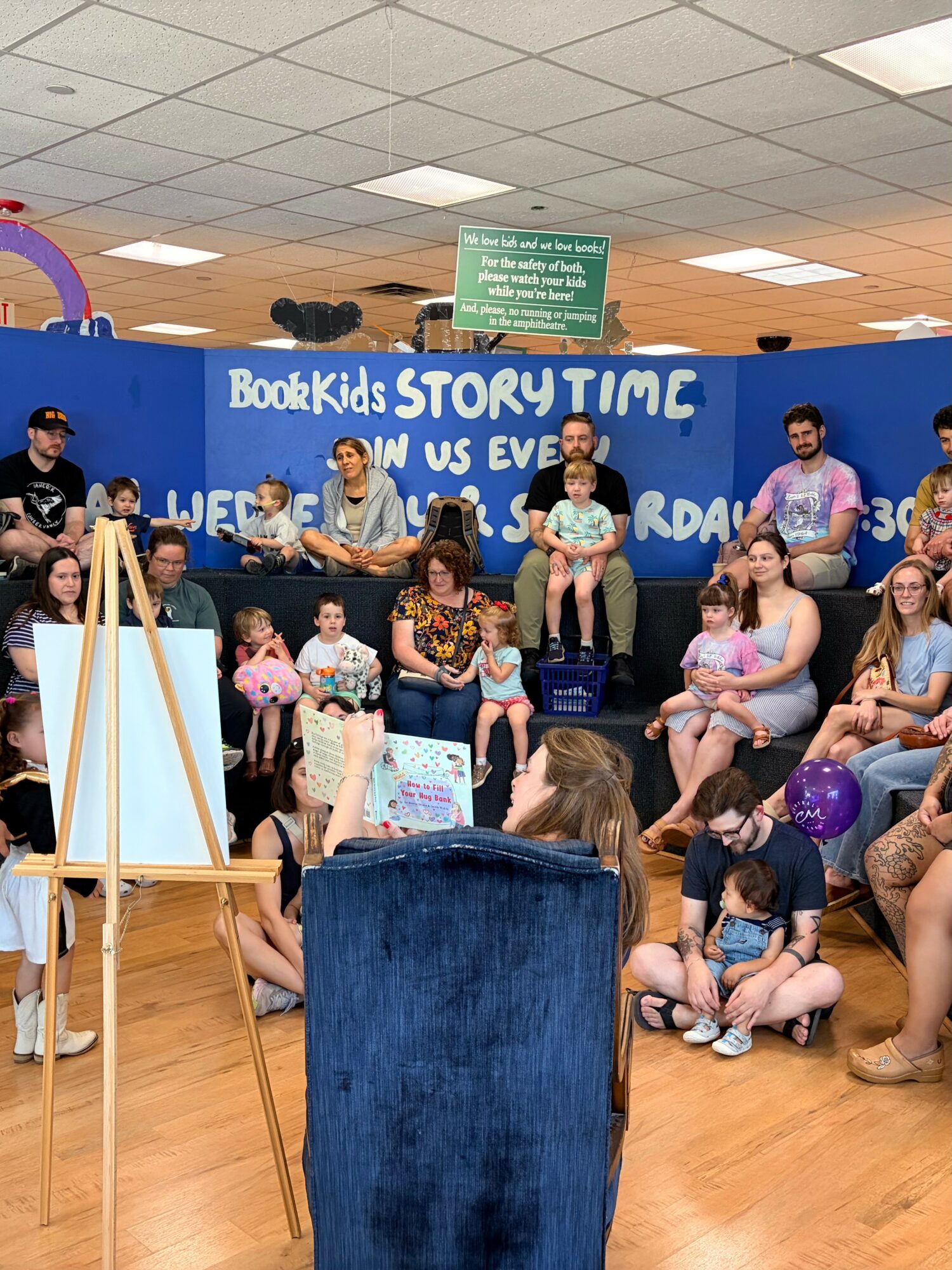 Children and adults gather on stage for a storytelling event in a room with a blue backdrop and a sign reading 'BookKids STORYTIME.'