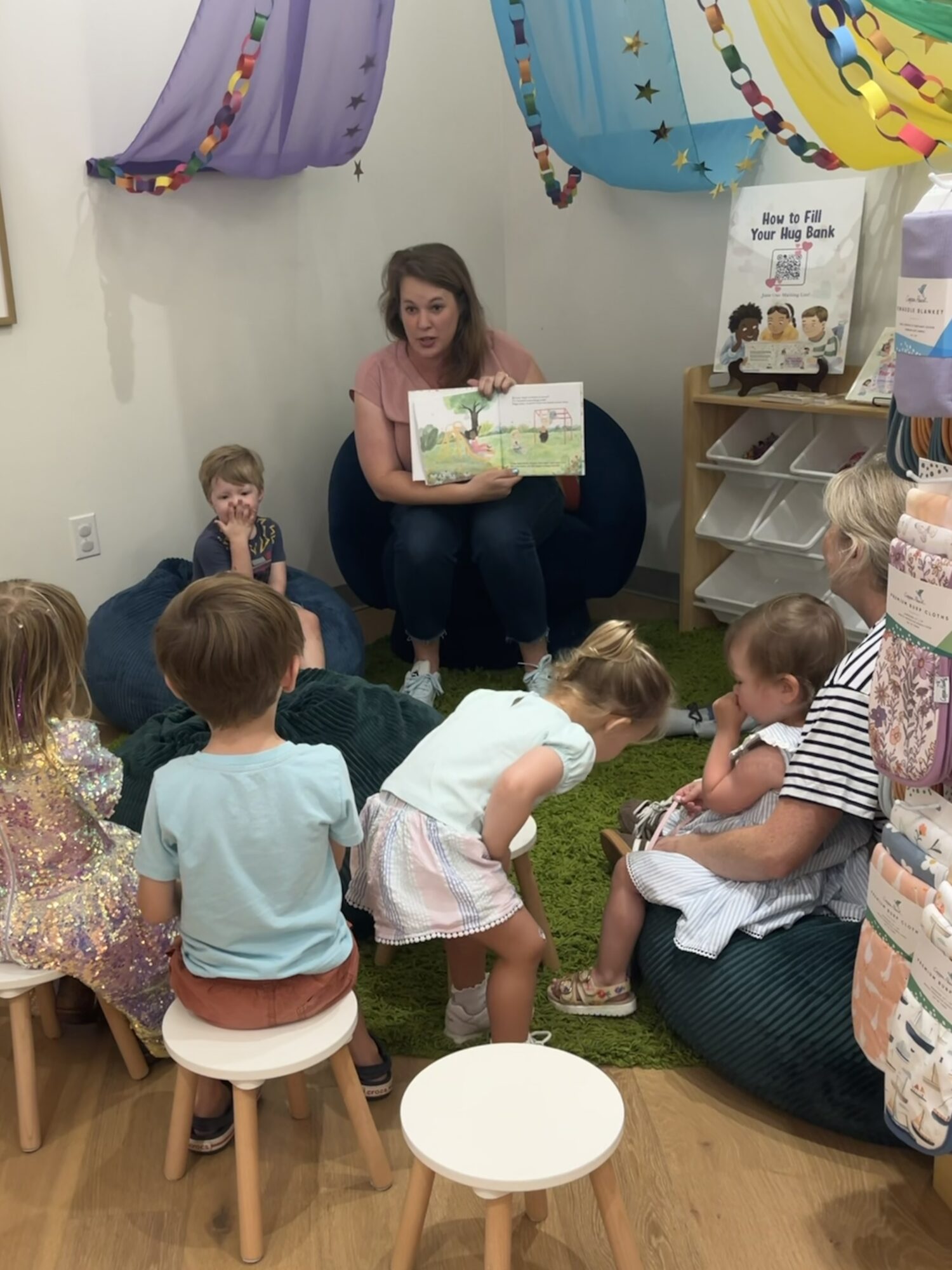 Woman reading a picture book to children seated on the floor in a colorful room with hanging decorations.