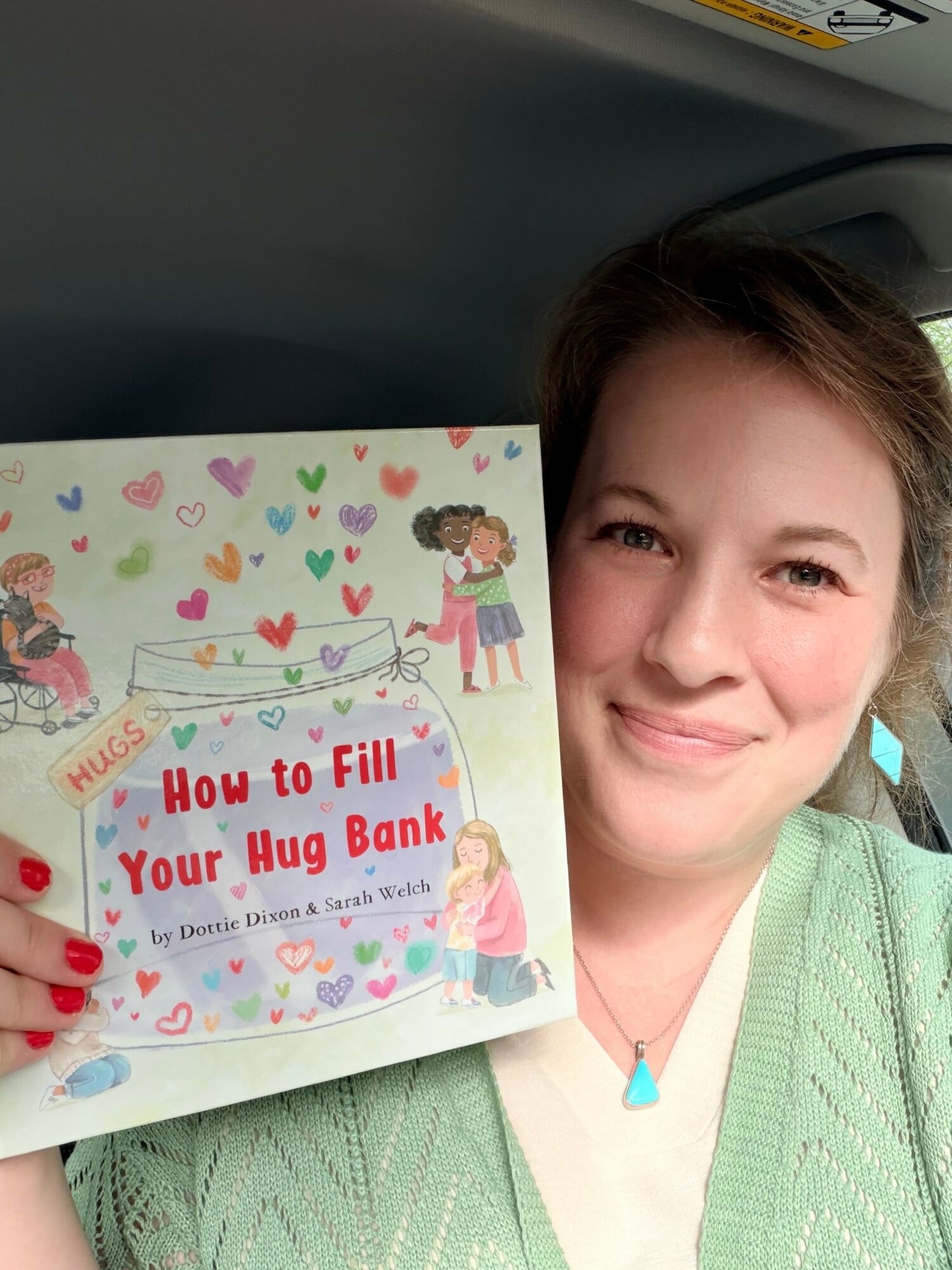 Woman holding a colorful book titled 'How to Fill Your Hug Bank' with illustrations of children and hearts, smiling.