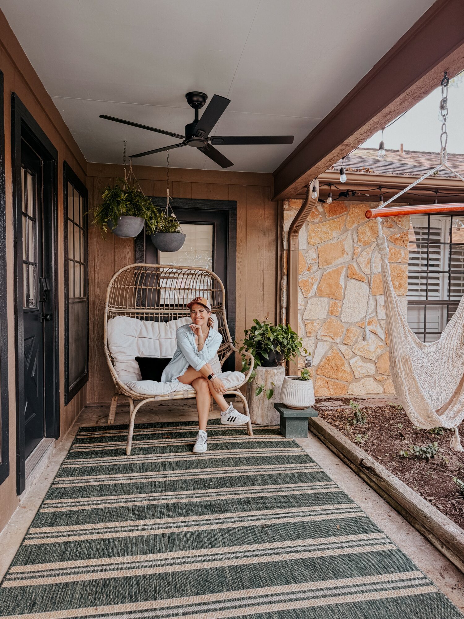 Porch with wicker bench, hanging plants, potted plant, and hammock, with stone wall and window, striped rug, ceiling fan, and porch ceiling.