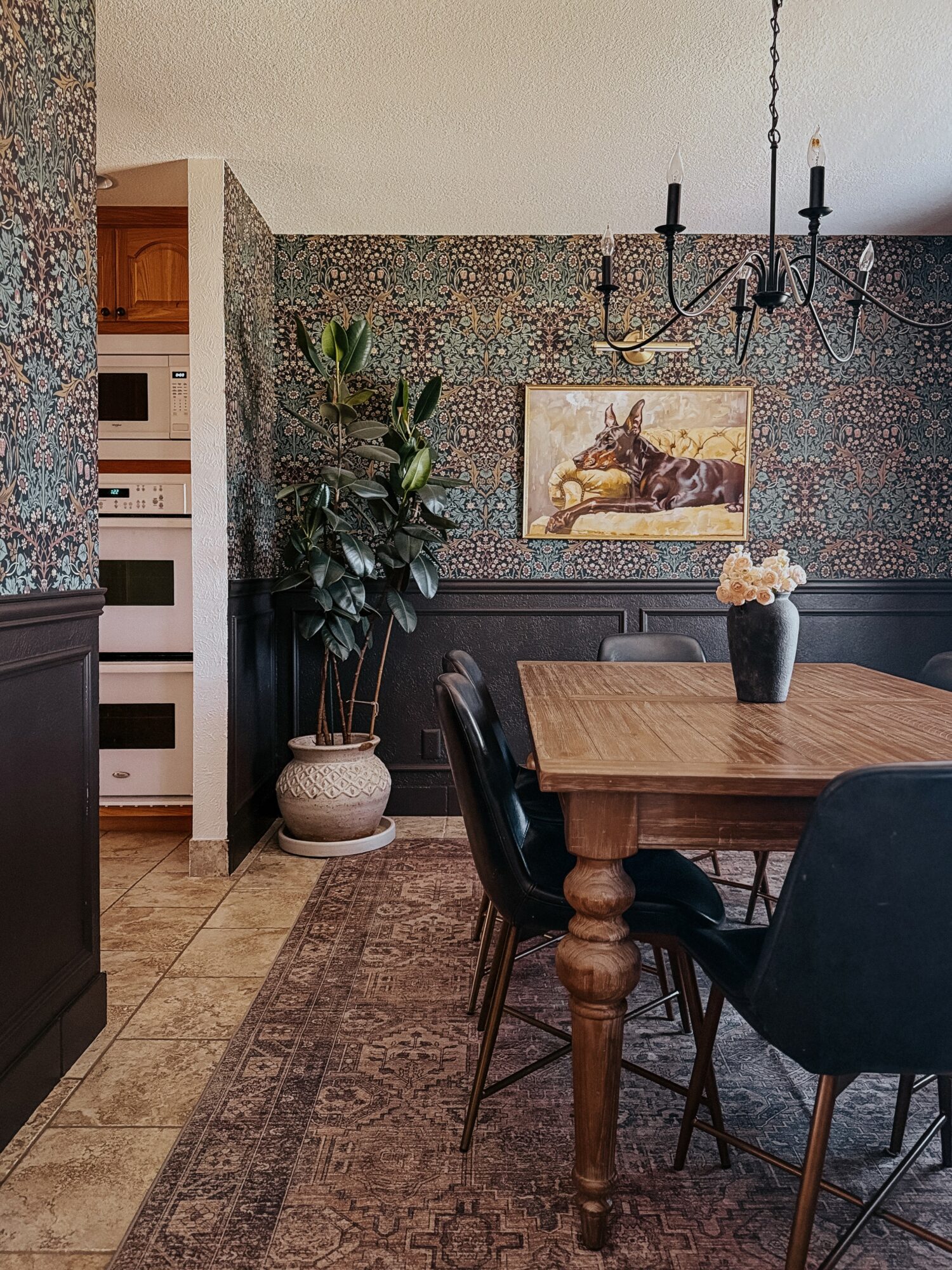 Dining room with wooden table, black chairs, large plant, painting, chandelier, and patterned wallpaper.