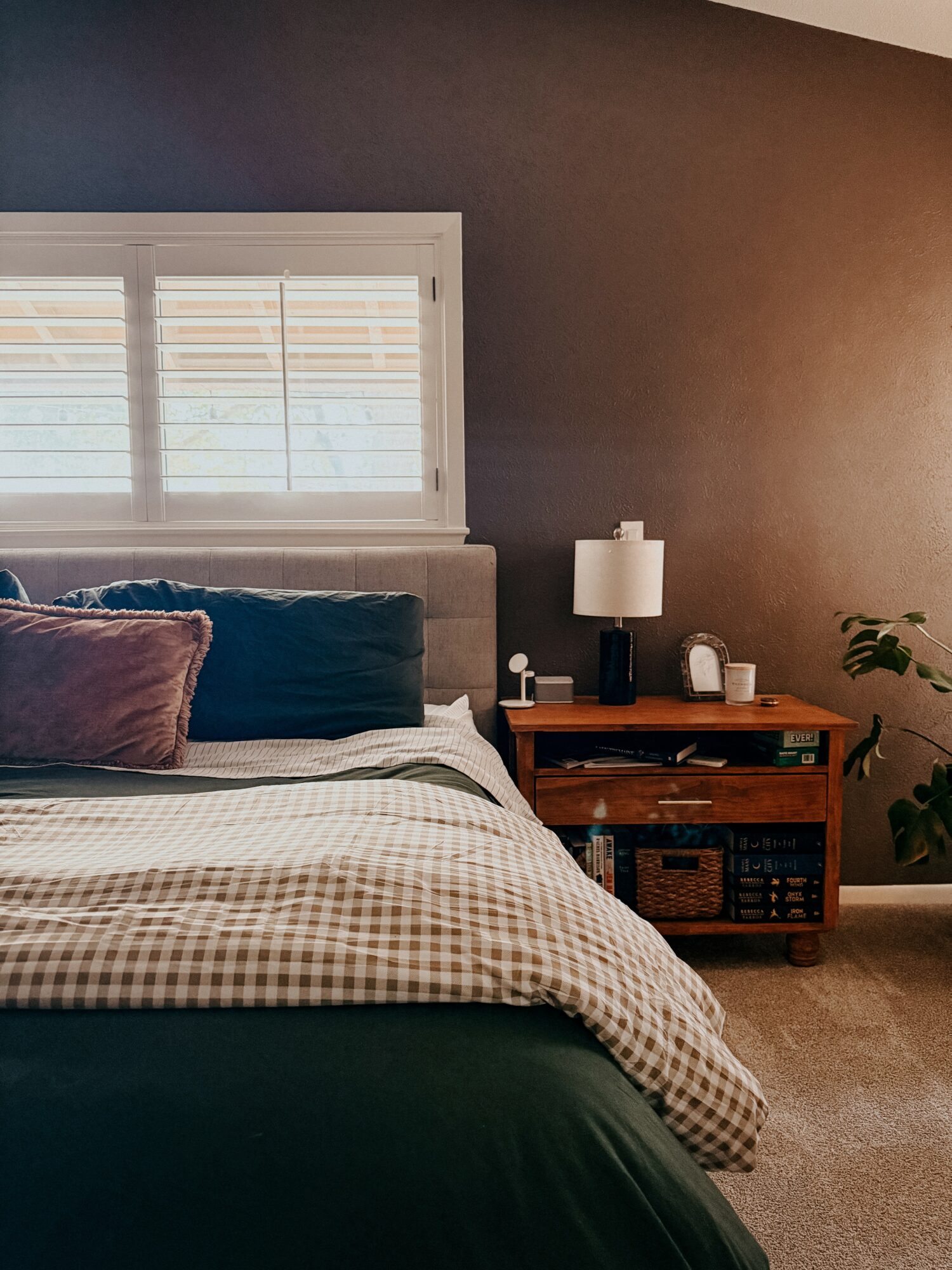 Bedroom with window, bed, nightstand, lamp, and plant, in warm lighting.