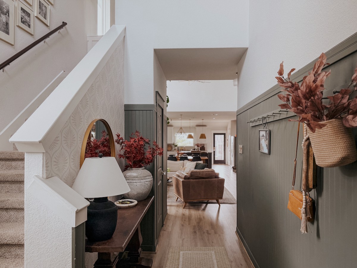 Hallway leading to living room with furniture and decorations, wall with hanging flowers on the right, and a table with a lamp on the left.