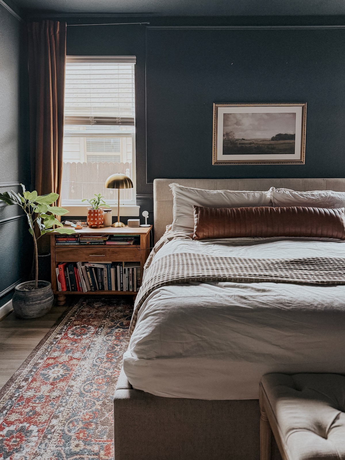 Bedroom with dark blue walls, a window with blinds, a bed with pillows, a wooden nightstand with books, a lamp, and a framed picture.