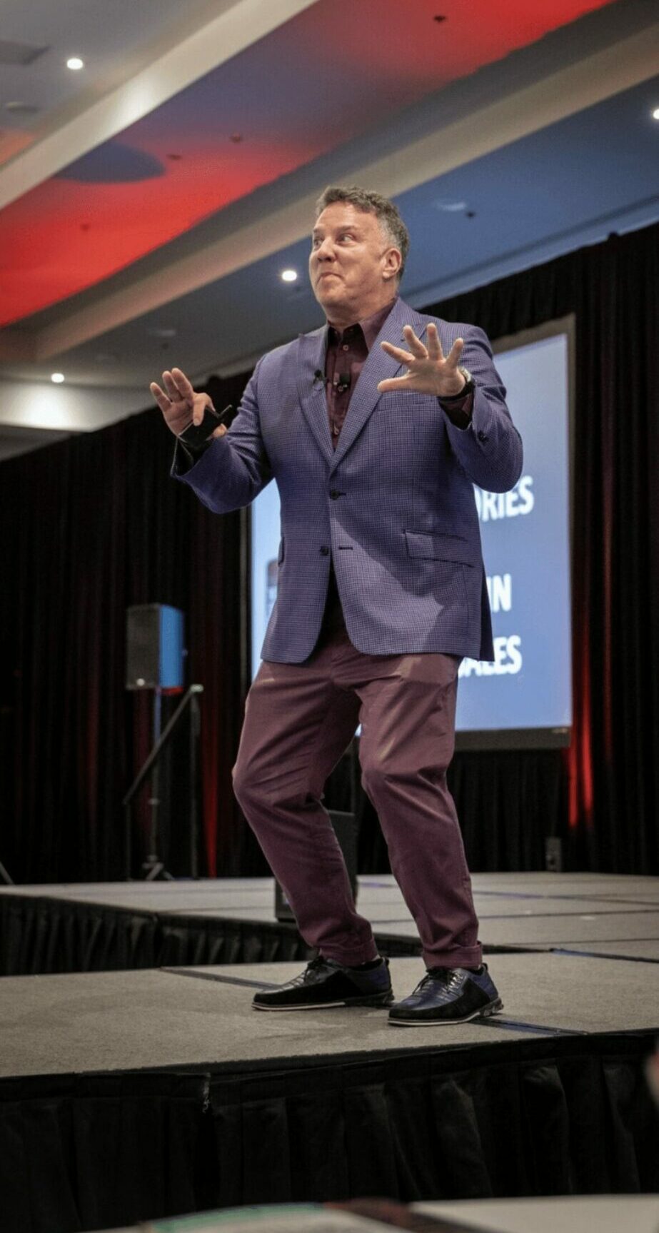 Person on stage speaking with hands raised, wearing a blue blazer and maroon pants, in a conference room.