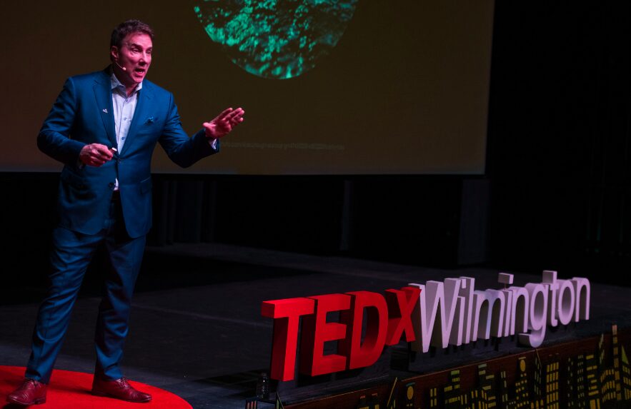 Man in a suit speaking on stage with TEDxWilmington sign, dark background, presentation screen behind him.