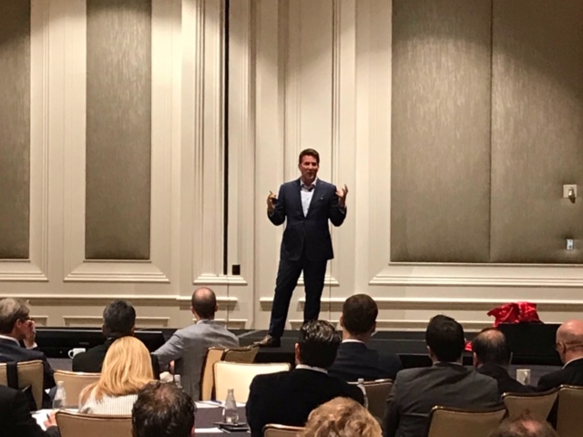 Man in a suit speaking on stage in front of an audience in a conference room.