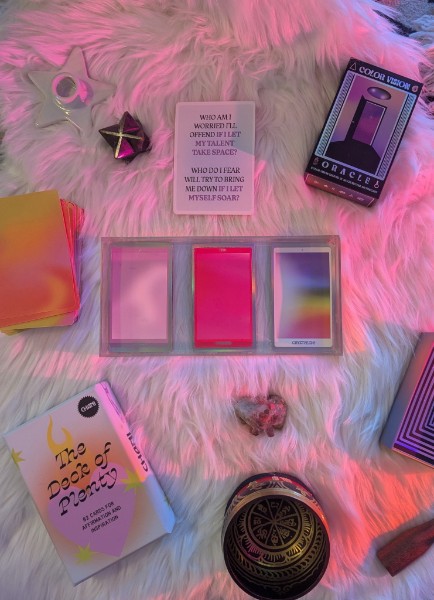 Pink fluffy surface with tarot cards, a box, a small heart, and various colorful objects, including a book and a round container.