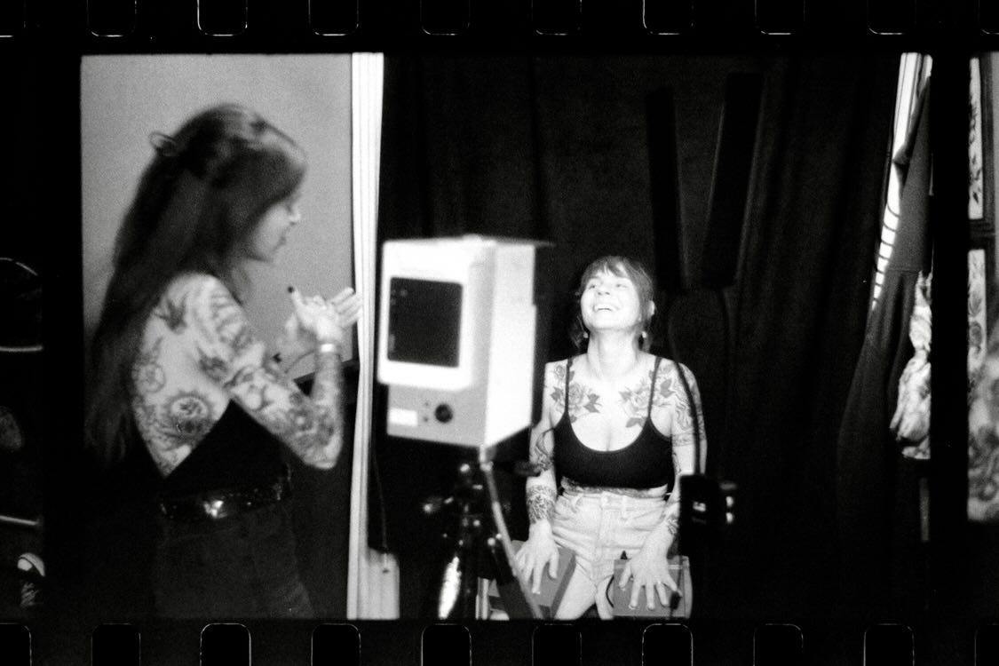 Two women in a room, one with long hair and patterned clothing, the other with short hair and a camera, smiling.