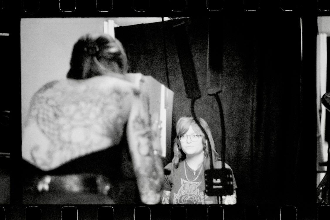A person with tattoos and a tattooed back faces a woman with glasses, in a dark room with curtains.
