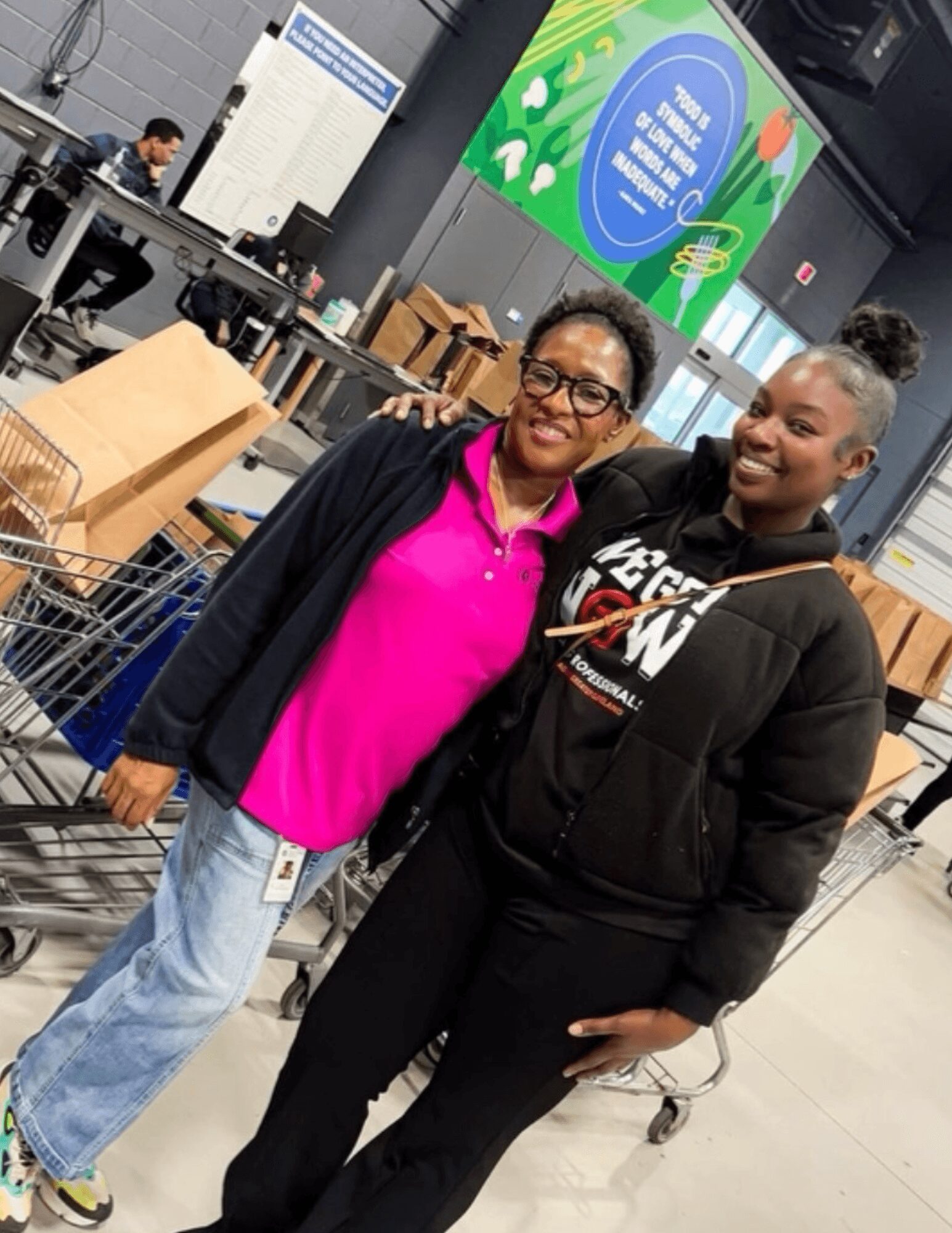 Two women smiling in a store with shopping carts and boxes, one wearing a pink shirt and glasses, the other in black hoodie.
