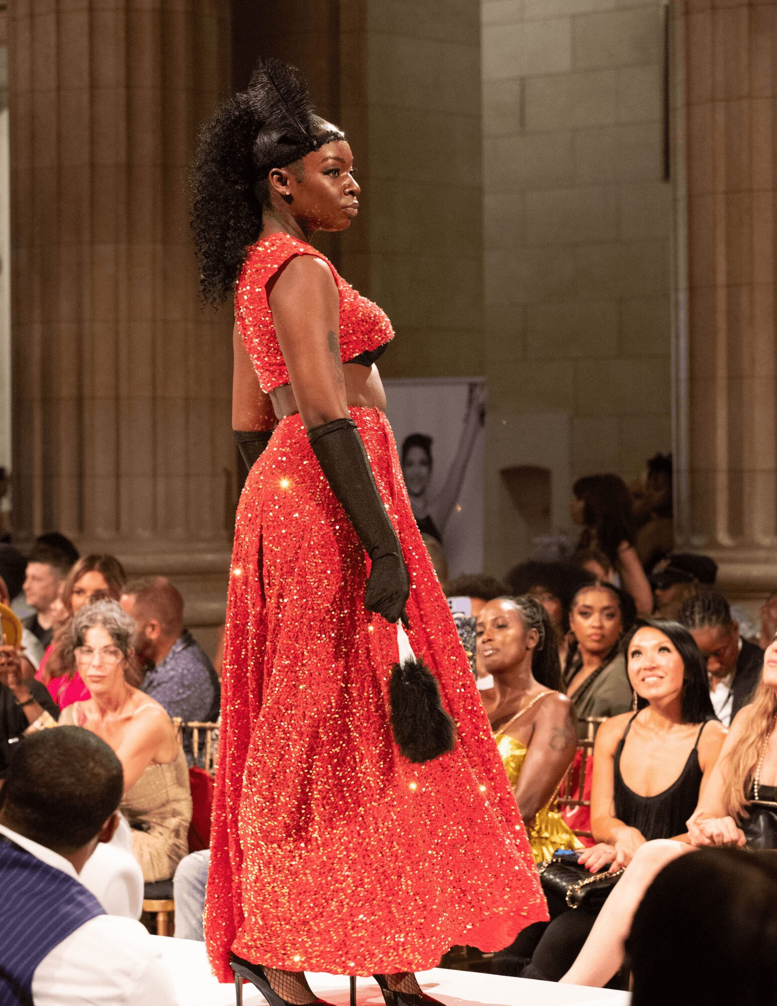 Model in red dress walking on runway with audience in background.