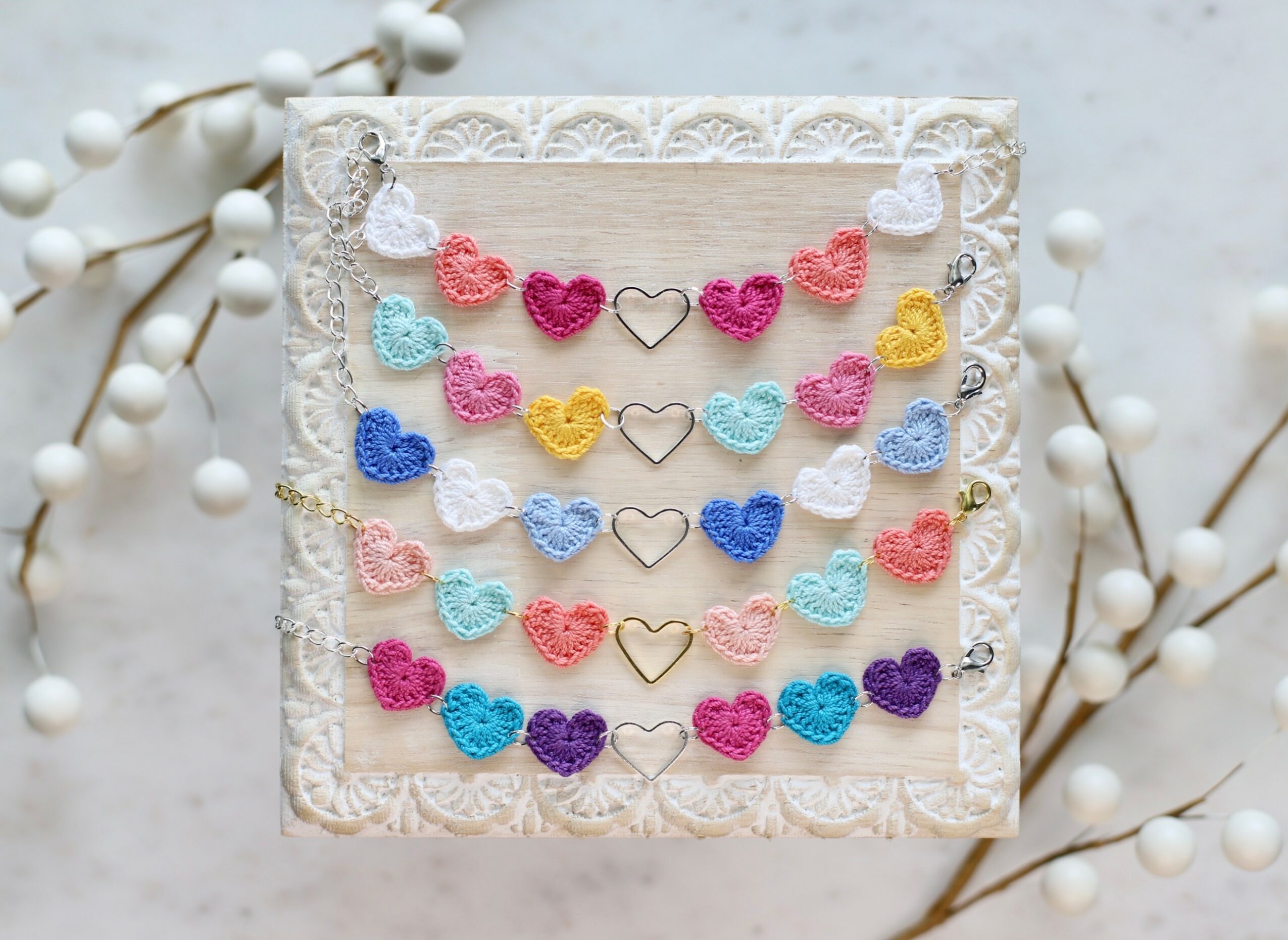 Square card with colorful crocheted hearts arranged in rows, surrounded by lace, on a white background with white decorative branches.