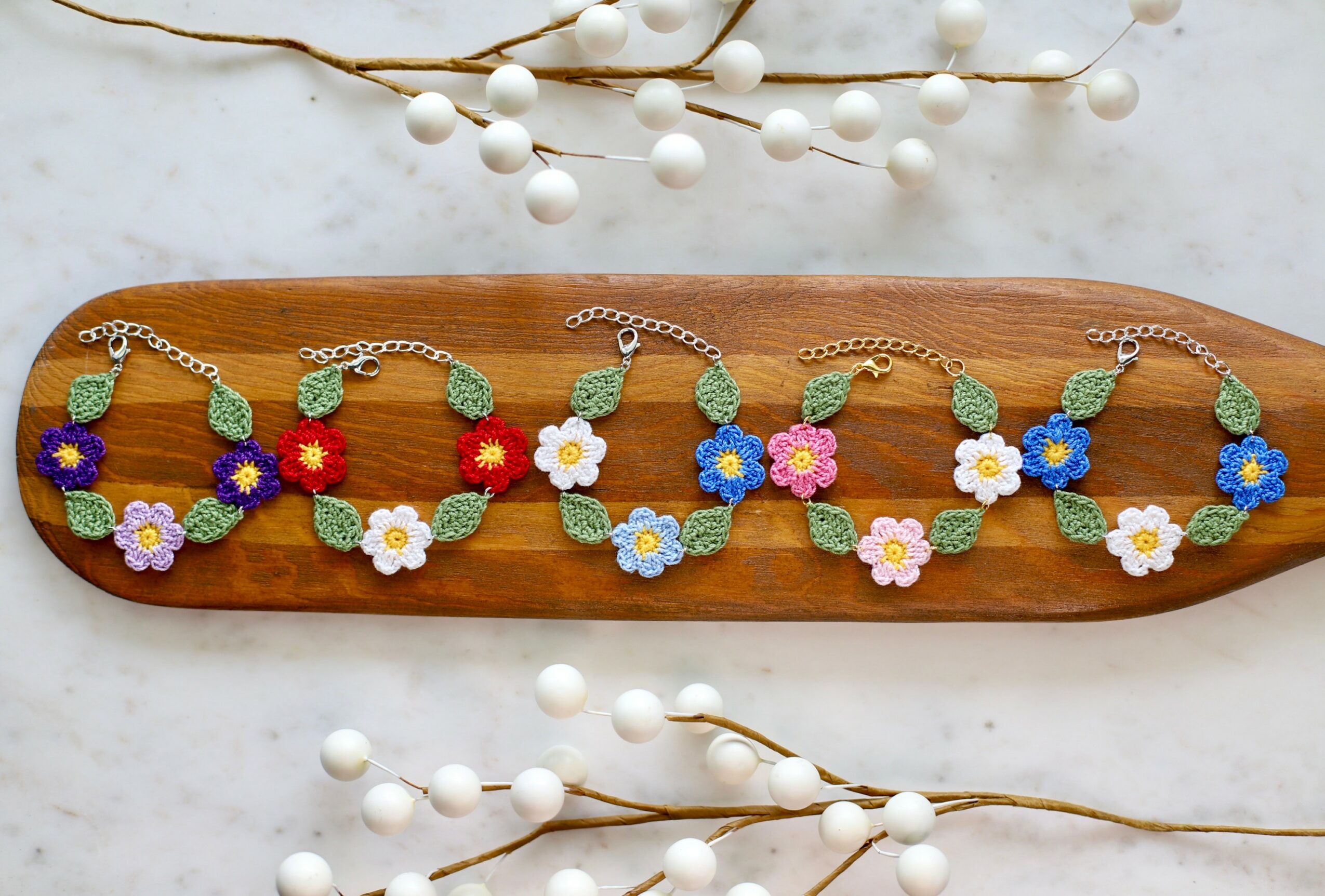 Colorful floral bracelets with leaves and chain links displayed on a wooden board, with white decorative berries in the background.