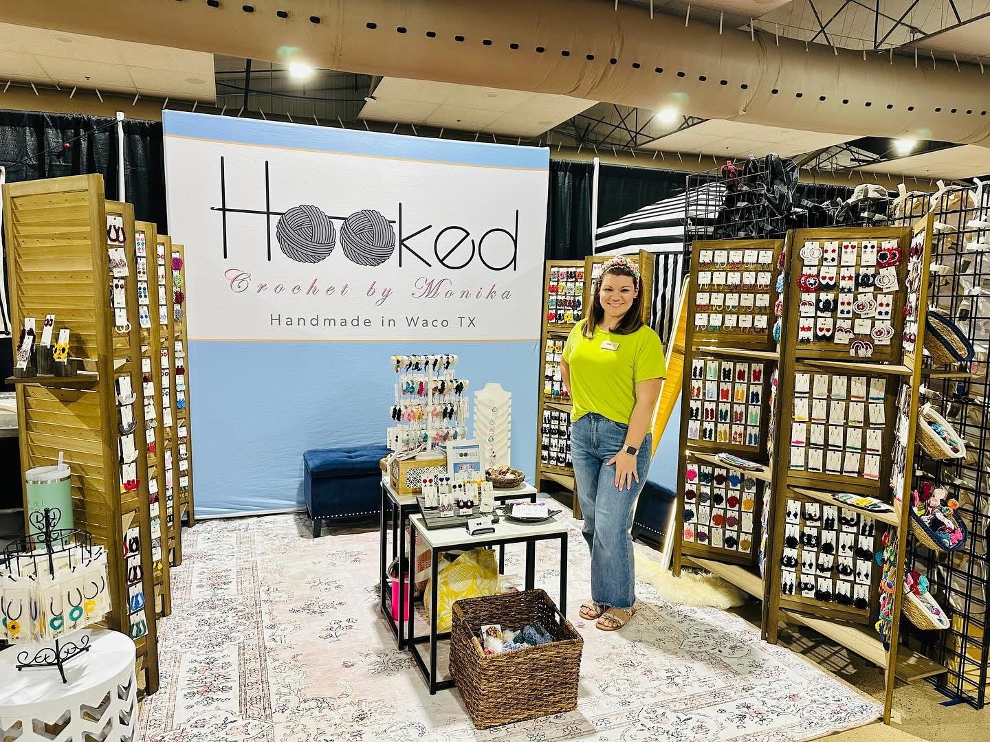 Woman standing in a craft booth with display shelves and a sign that reads 'Tucked, Handmade in Waco TX'.