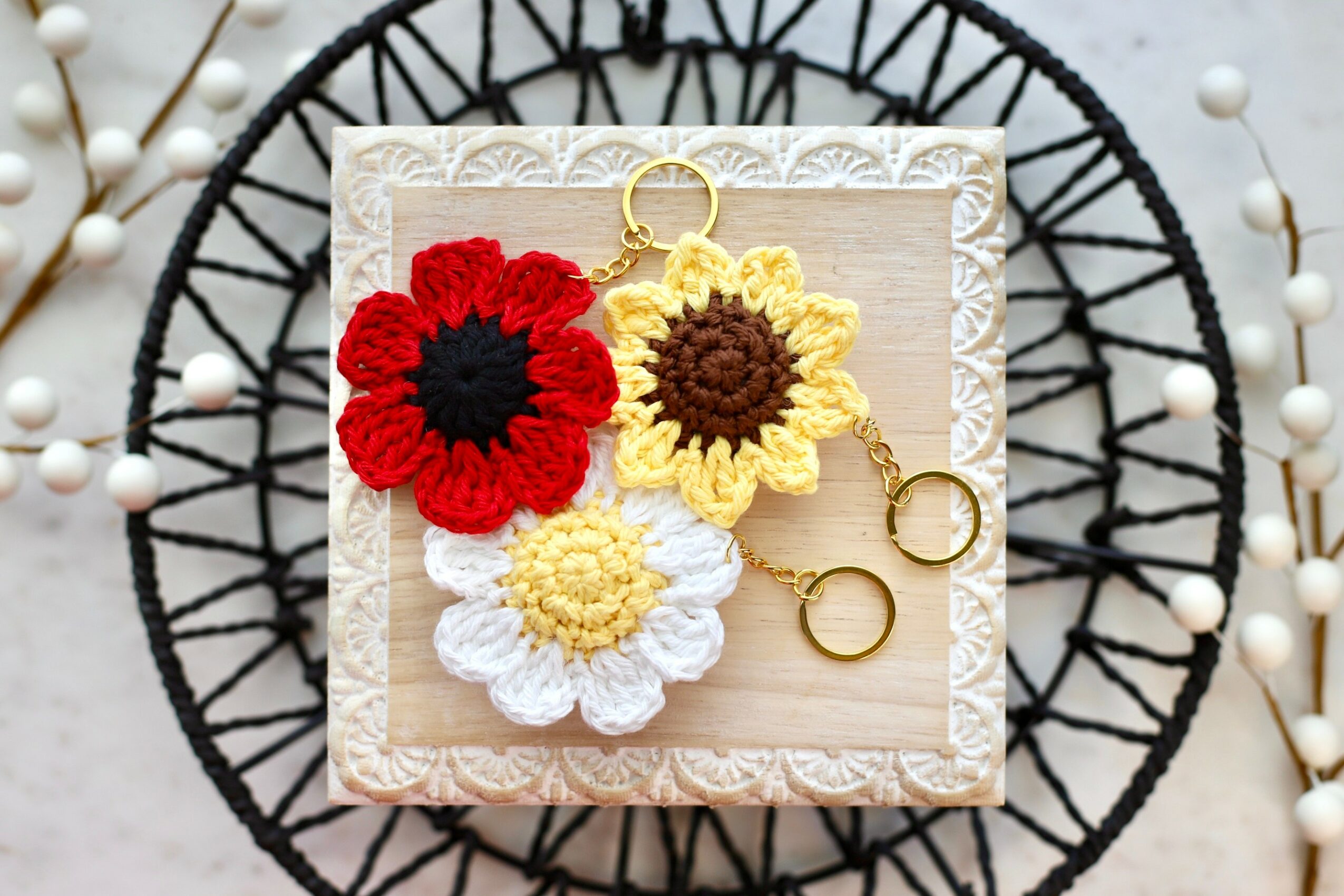 Three crocheted flower keychains in red, yellow, and white on a decorative square frame, with a black circular wire background.