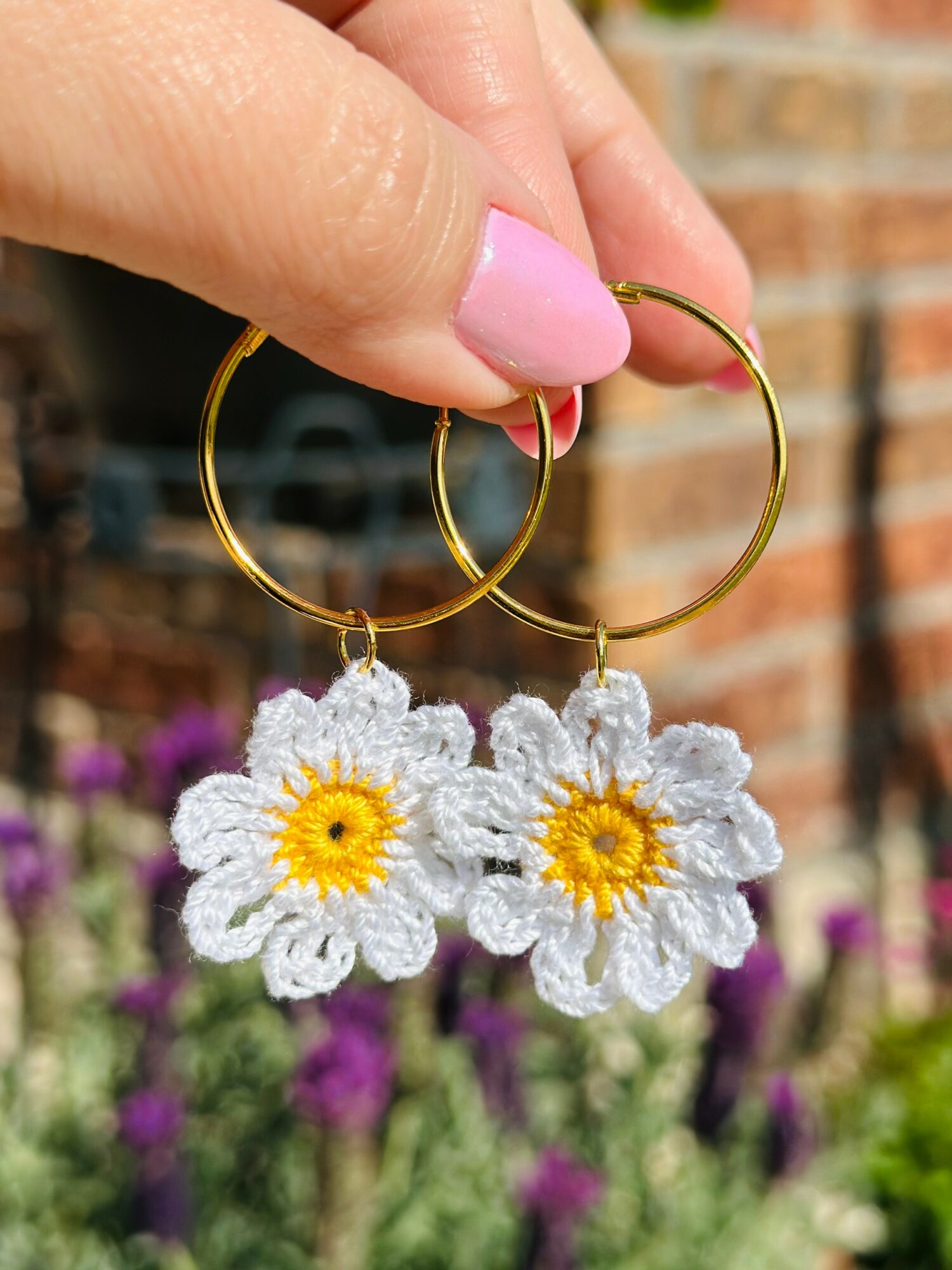 Hand holding gold hoop earrings with white daisy flower charms, outdoor background with brick wall and purple flowers