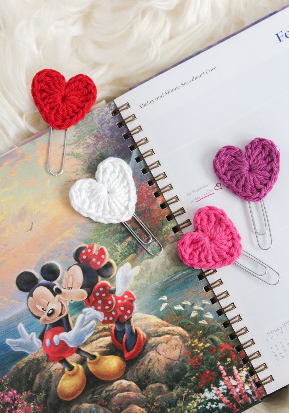 Open notebook with colorful illustrations, three knitted hearts in red, white, pink, and purple, and paper clips on a soft surface.