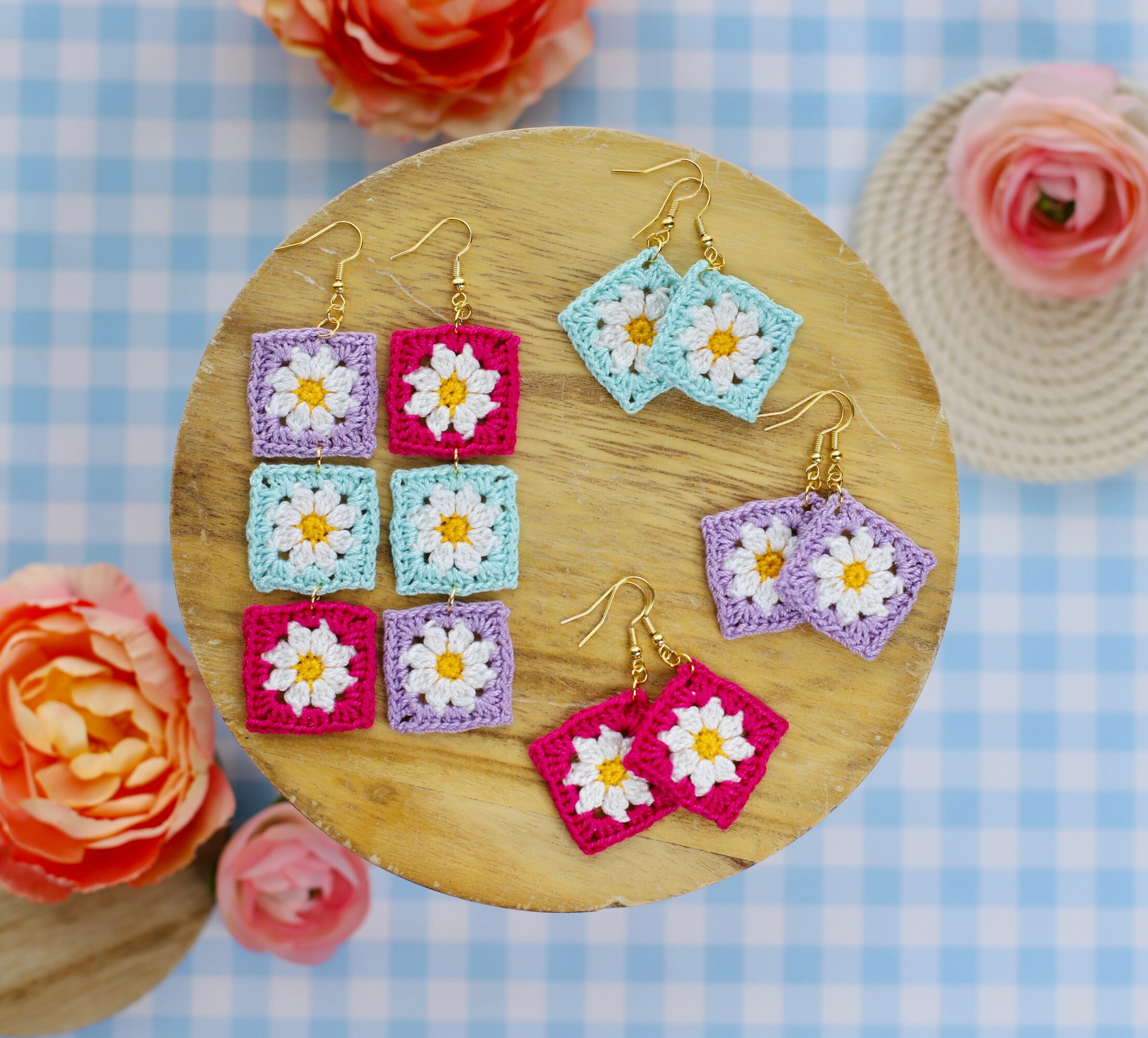 Colorful crochet earrings with daisy flower designs on a round wooden surface, surrounded by pink and orange flowers, on a blue checkered background.