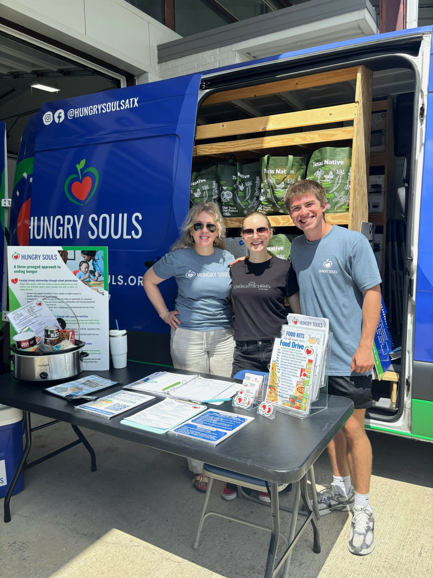 Three people stand in front of a blue mobile food truck with a table displaying informational materials, outdoors.