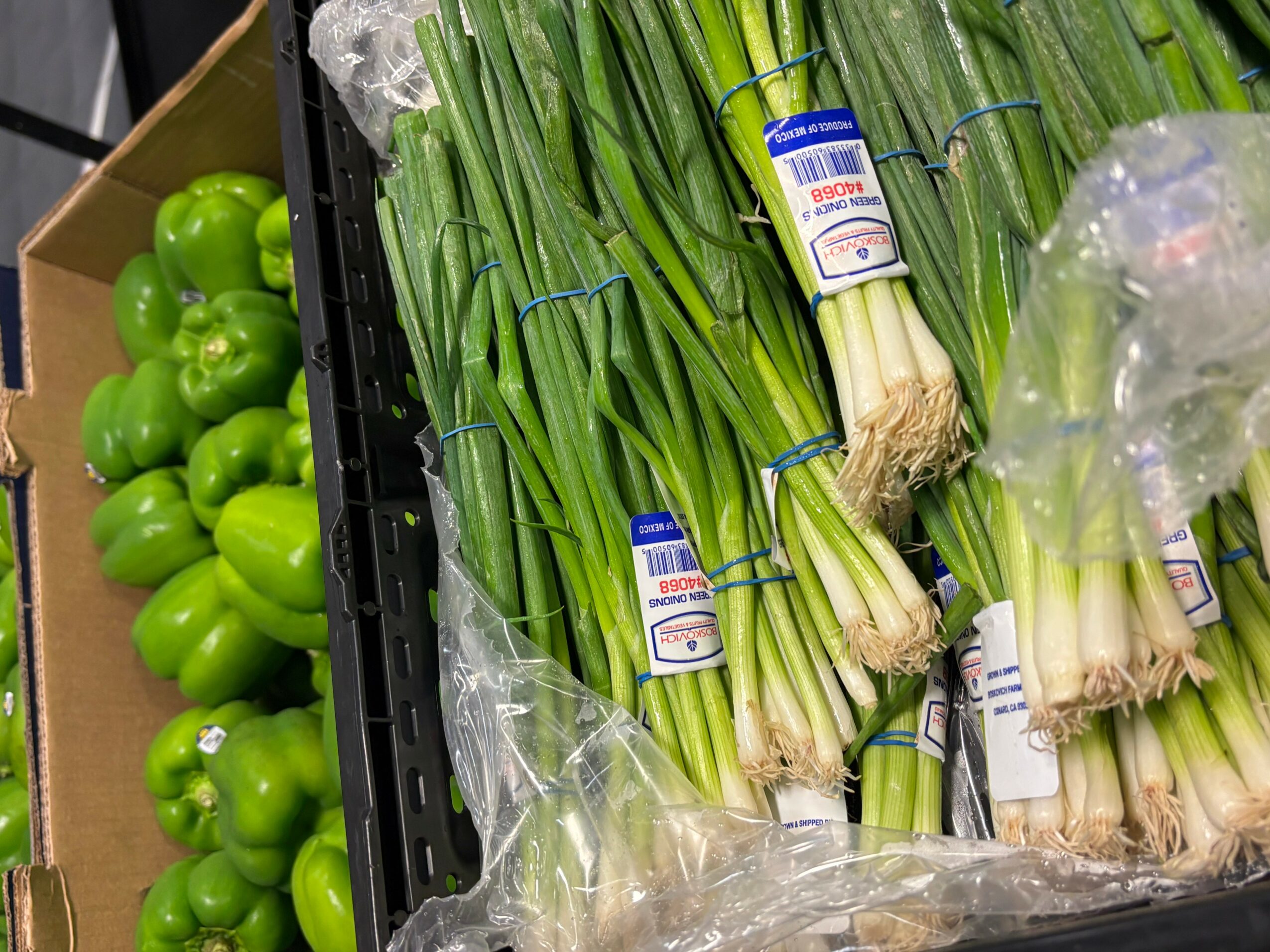 Green bell peppers in a cardboard box and bunches of green onions wrapped in plastic, displayed on a black tray.