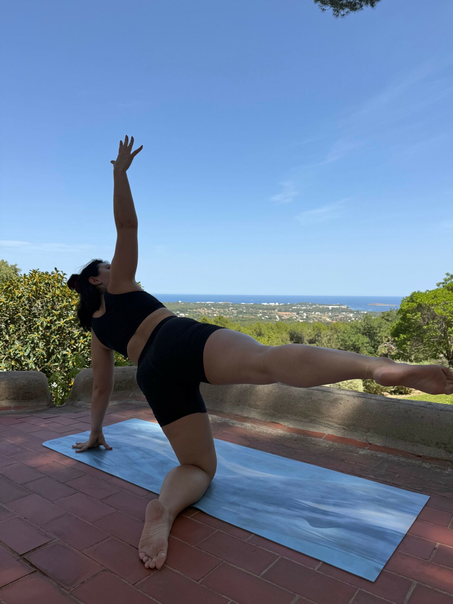 Person practicing yoga outdoors on a mat, with arms raised and one leg extended, against a background of trees and sky.