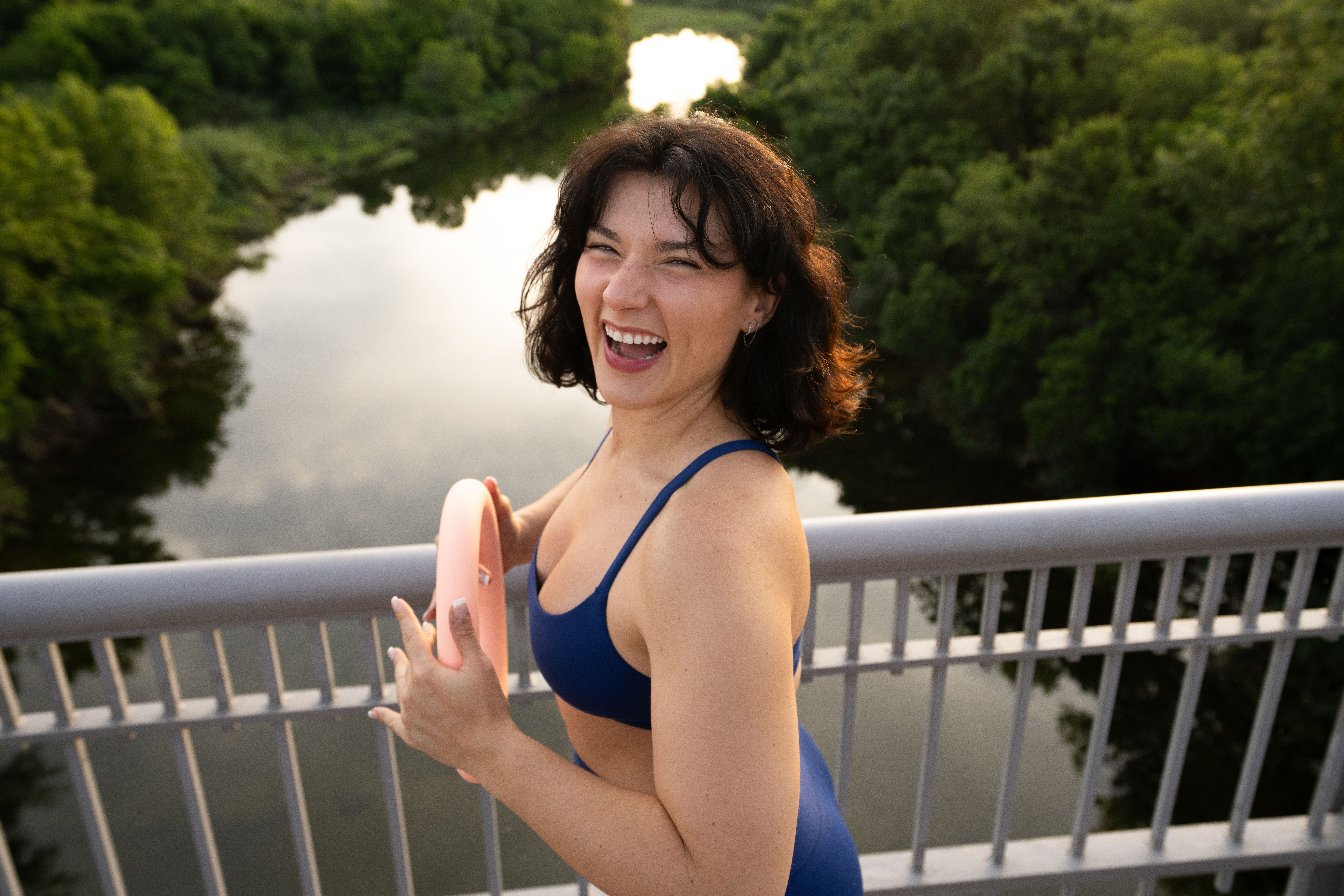 Woman in a blue swimsuit smiling and holding a pink object on a bridge over a river with trees.