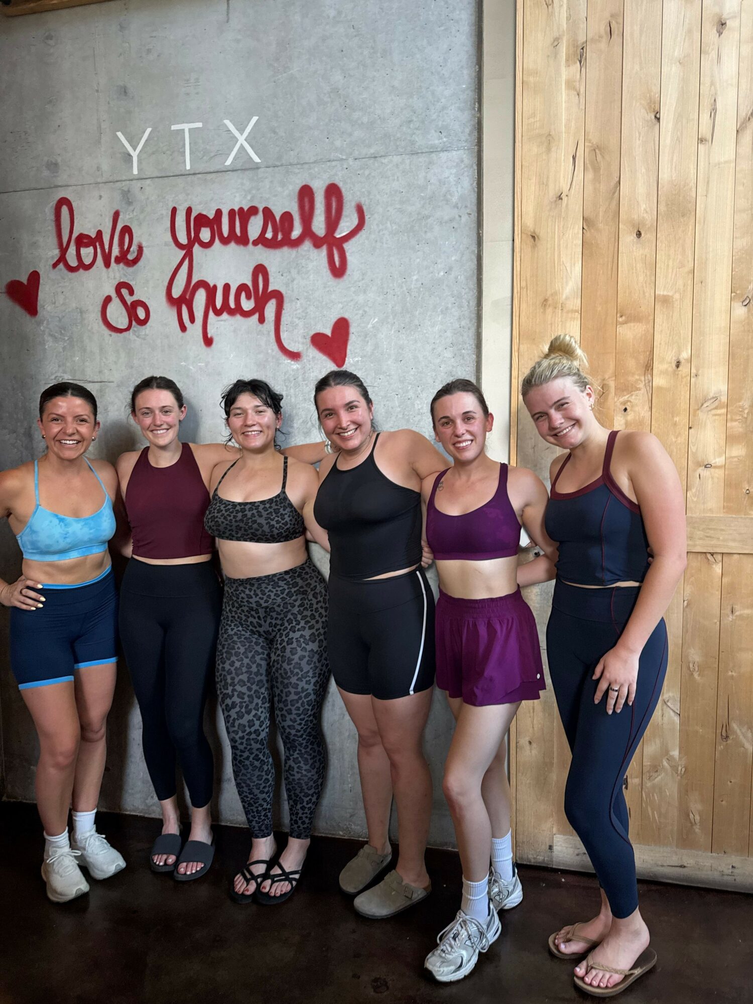 Group of six women standing together indoors, with a wooden wall and concrete wall behind them, smiling. Handwritten text says 'Love you and your' with a heart.