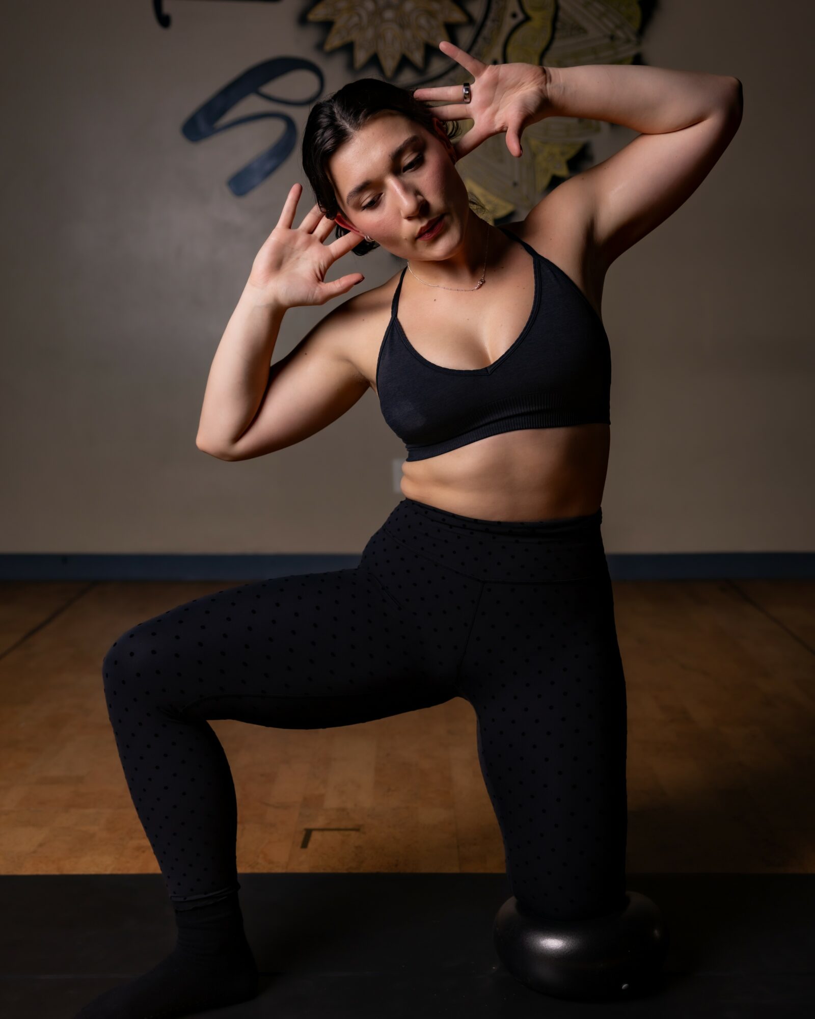 Young woman in workout clothes poses with hands near her face, one knee on a small exercise ball, in a gym.
