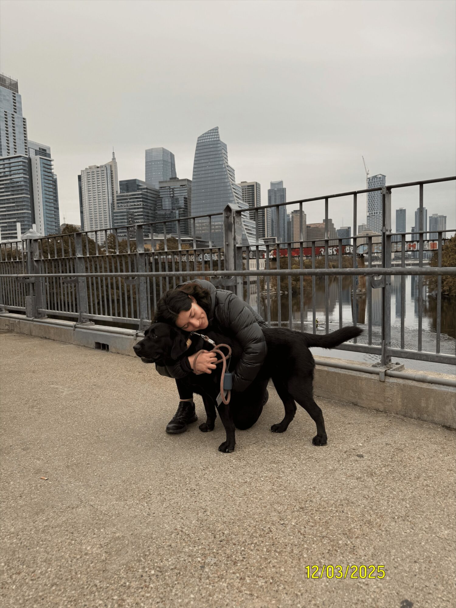 Two people hugging and petting a black dog on a city waterfront promenade with skyscrapers in the background.