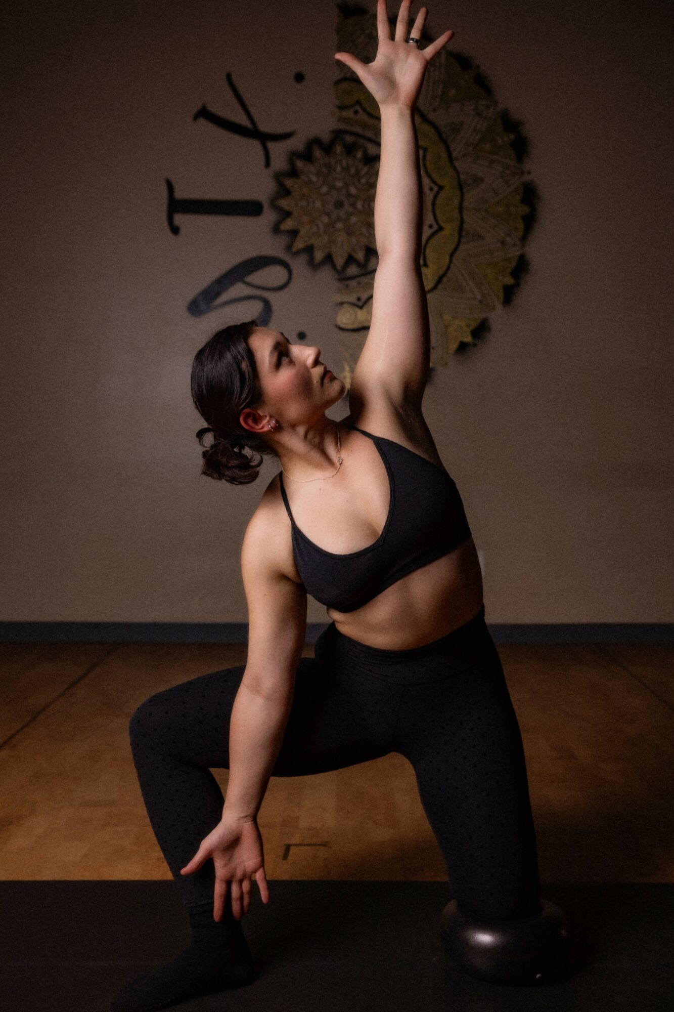 Woman in yoga pose with one arm raised, kneeling on one knee, in a studio with a decorative wall hanging.