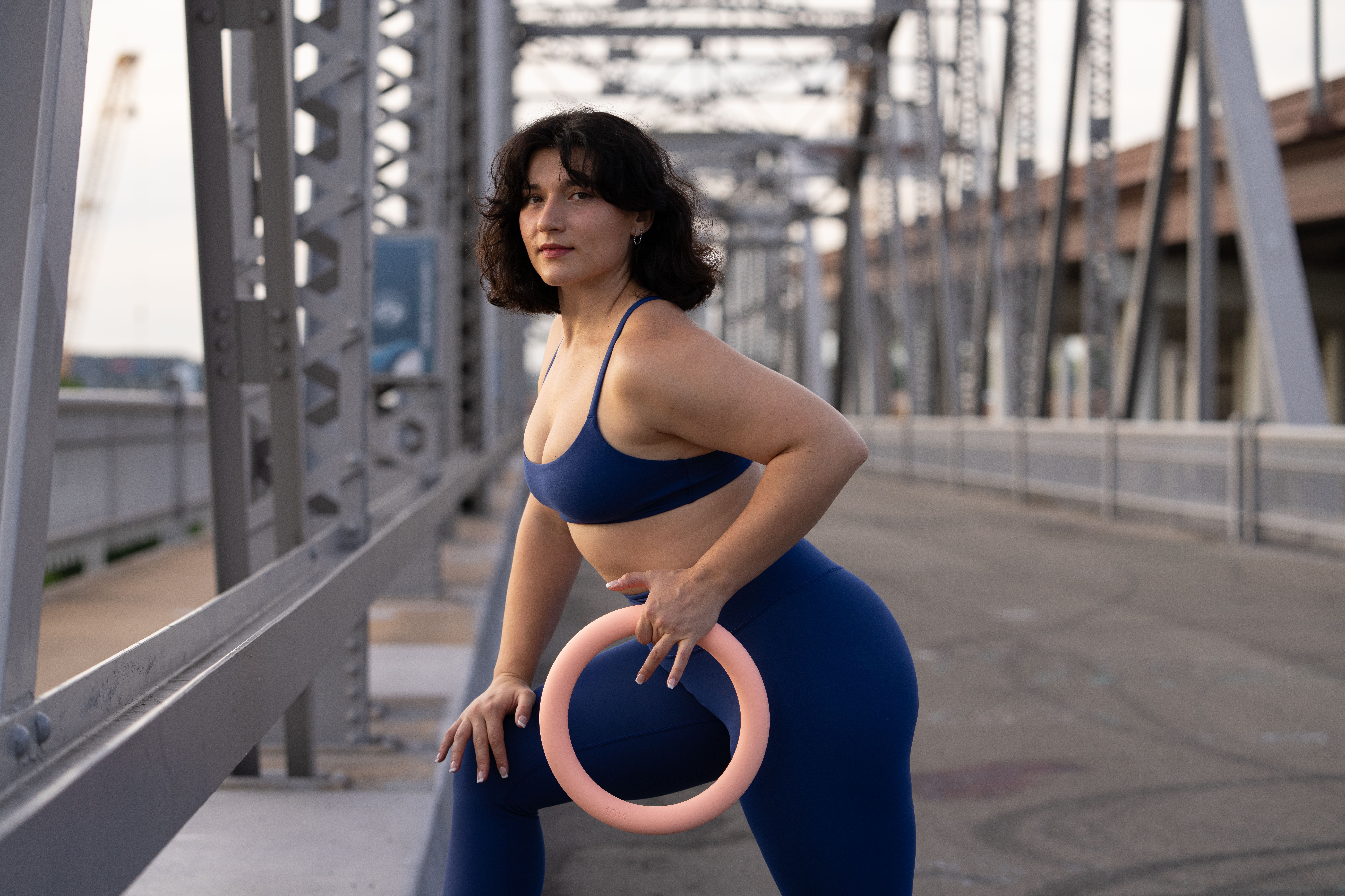 Woman in blue sportswear holding a pink ring on an outdoor bridge, with metal structures and cityscape background.