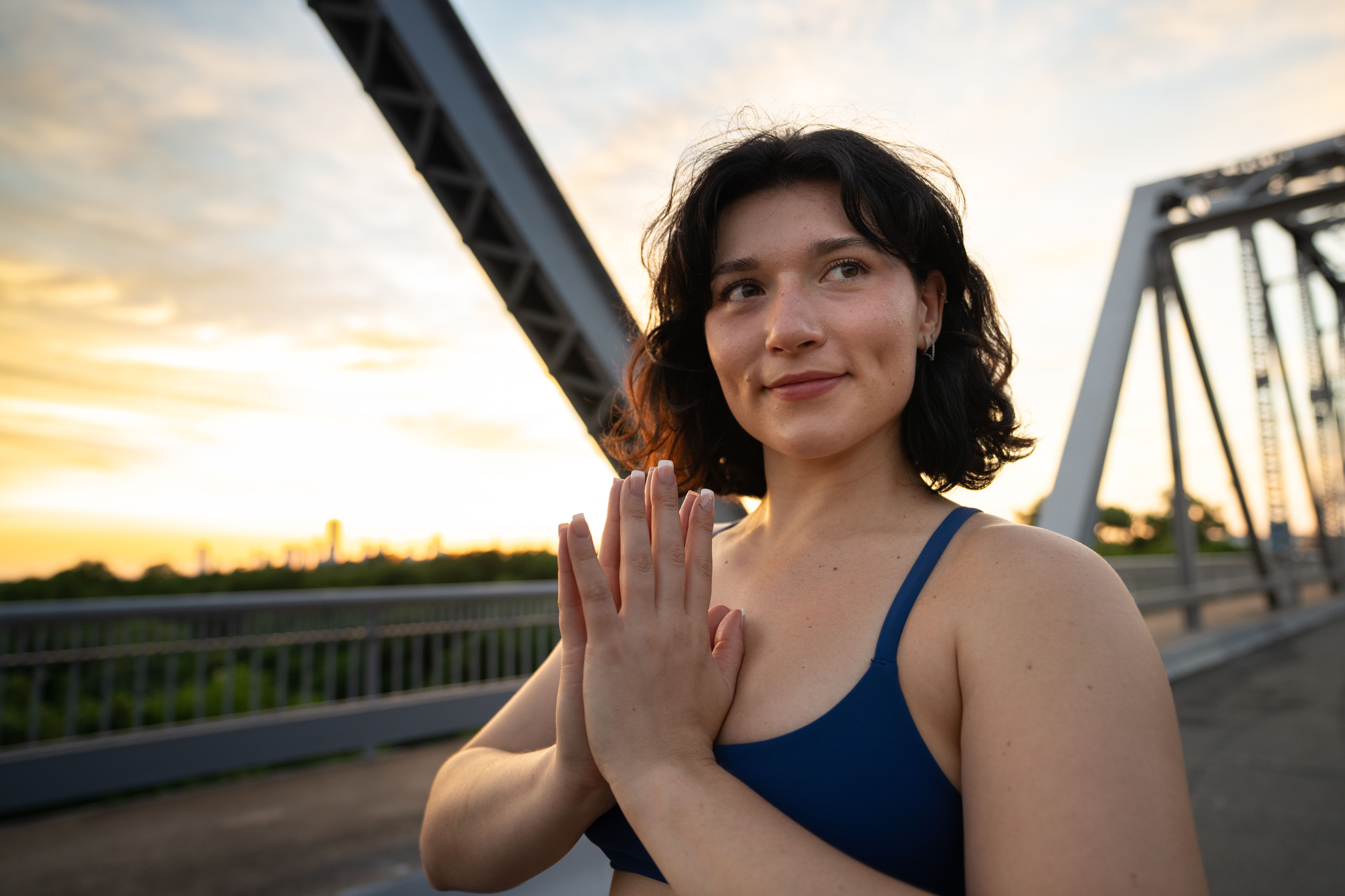 Young woman with dark hair in a blue tank top stands on a bridge at sunset, hands pressed together, smiling.