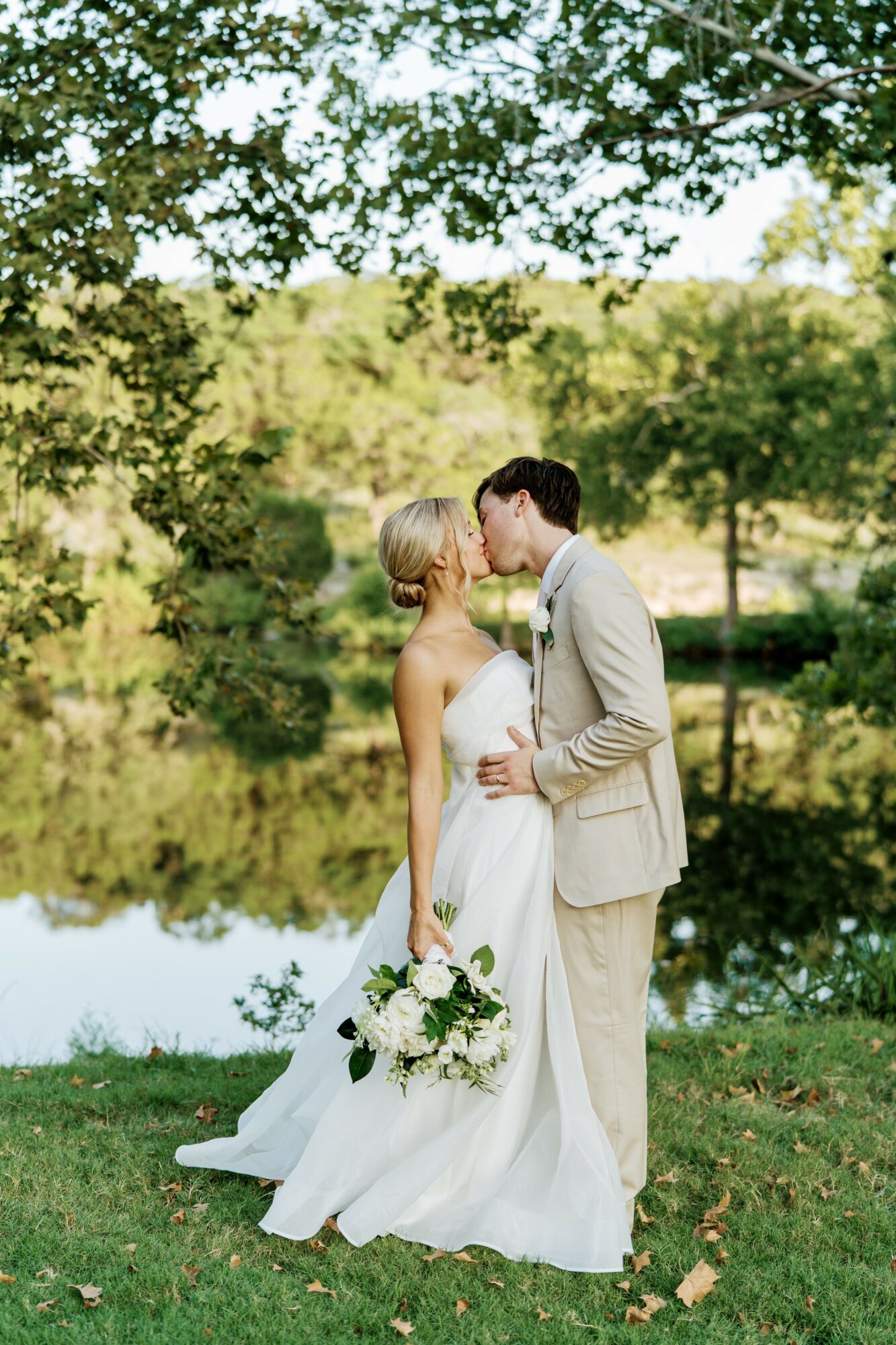 A bride and groom stand close by a lake with trees in the background, holding hands and gazing at each other.