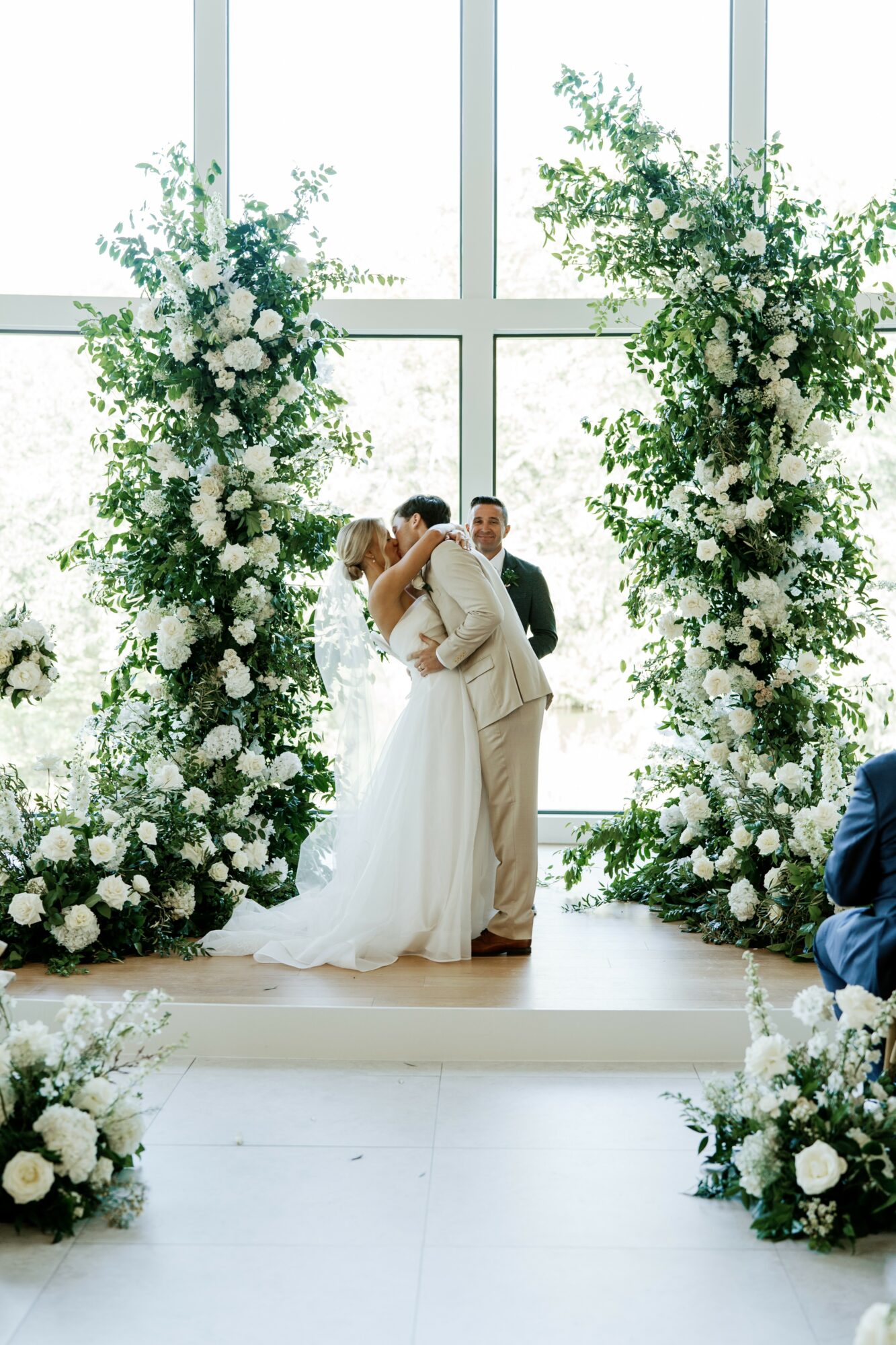 Bride and groom kiss at altar with floral arrangements and large window behind them, officiant in background, guests seated nearby.