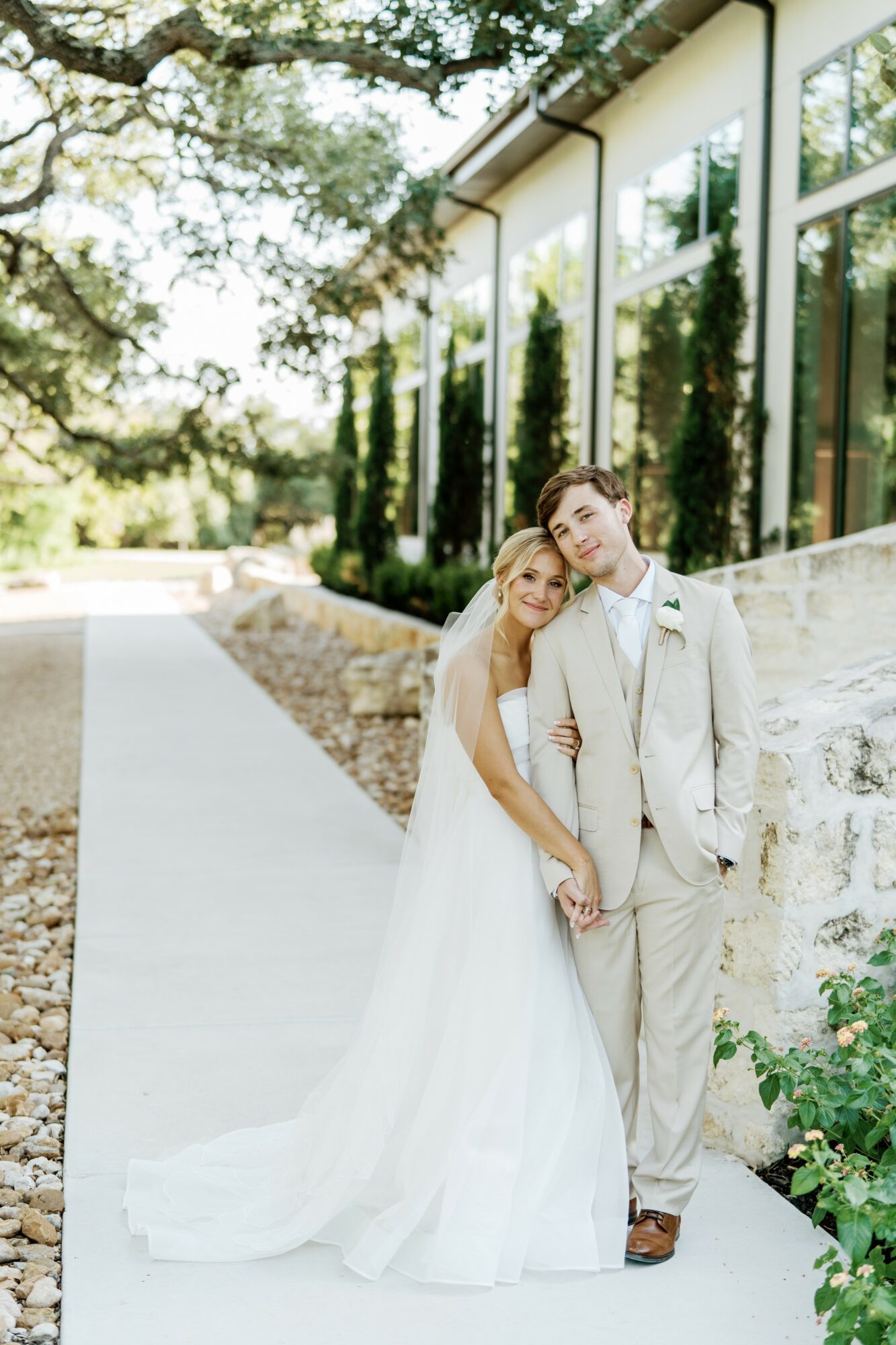 A bride and groom stand outdoors, holding hands, with a modern building and greenery in the background.