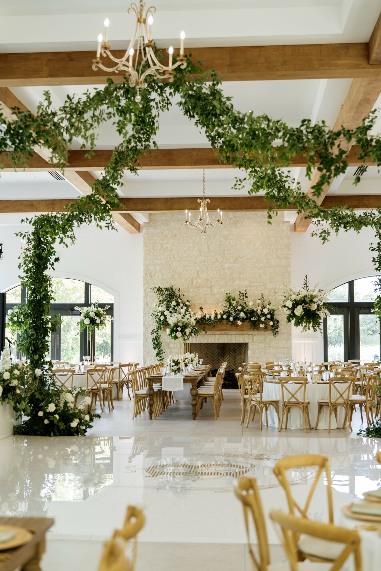 Elegant event space with greenery and chandeliers, decorated for a celebration with tables and chairs, large windows, and a fireplace.