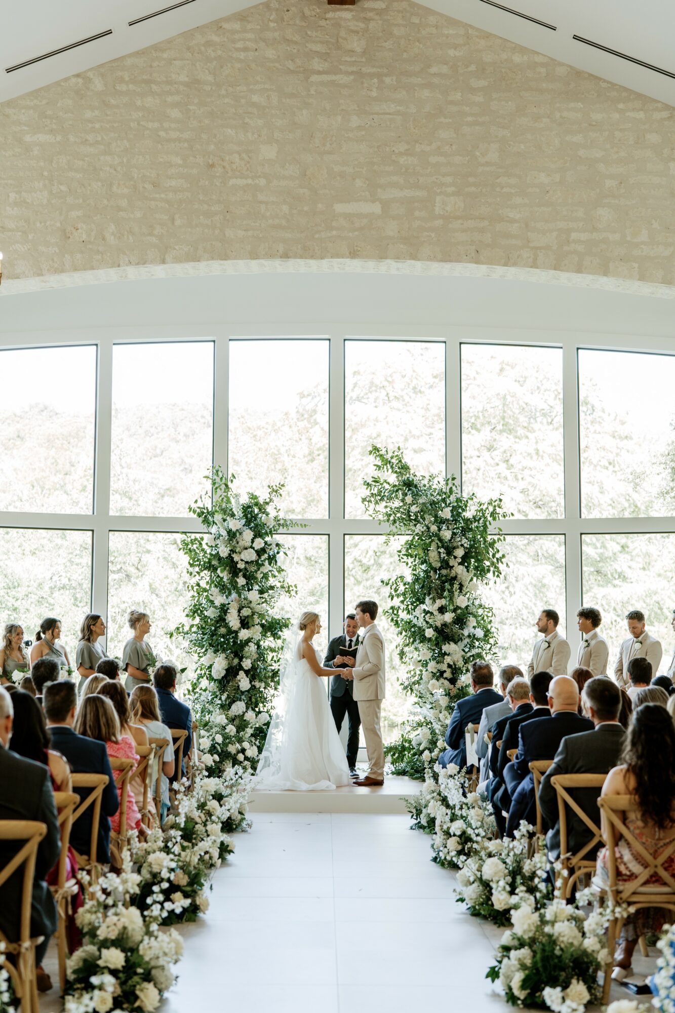 Wedding ceremony with bride and groom standing under floral arch, guests seated indoors, large windows in background.