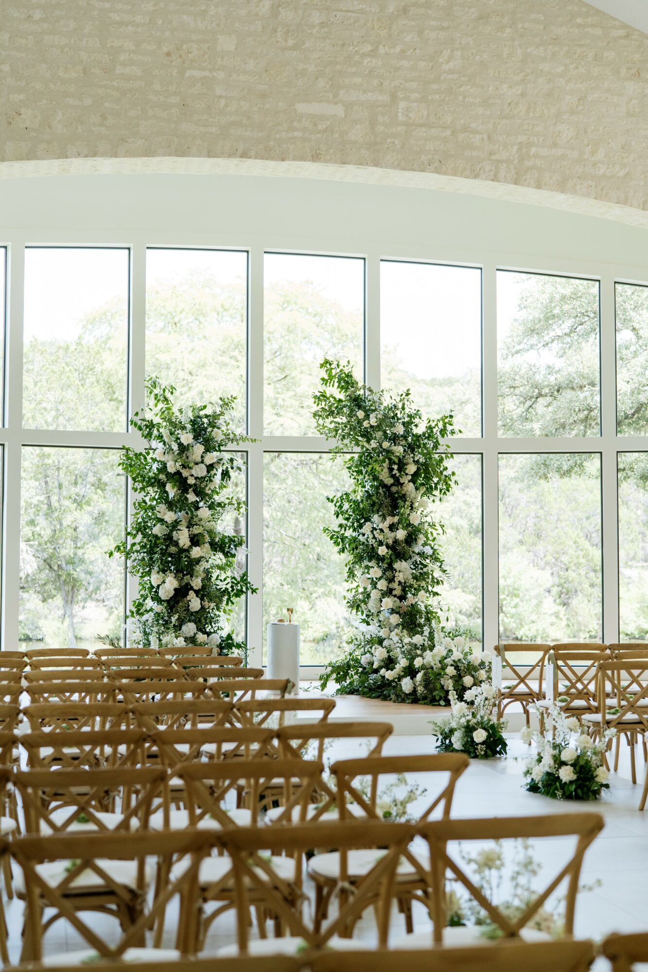 Interior of a bright room with large windows, decorated with floral arrangements and wooden chairs, likely for an event.