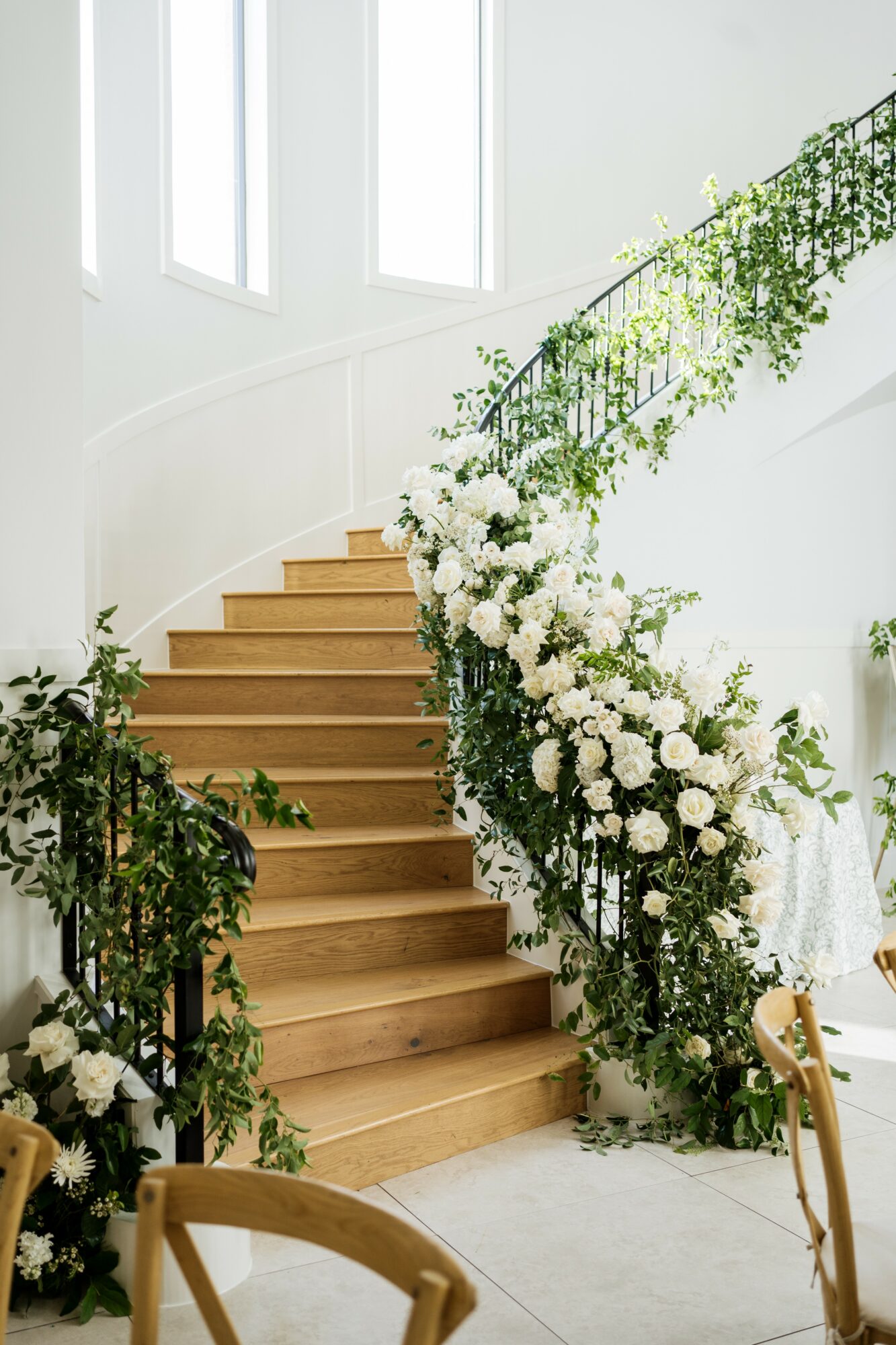 Wooden staircase decorated with white flowers and green foliage, inside a bright room with large windows.