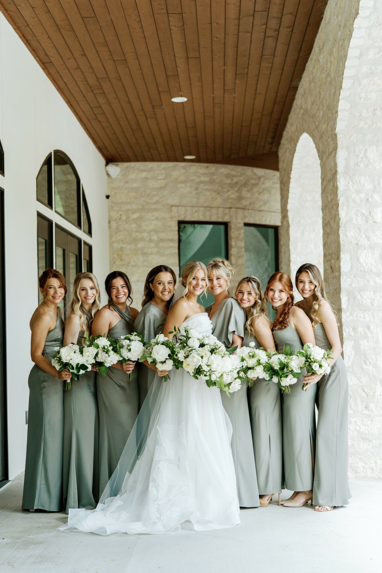 Group of ten women, including bride in white gown, holding large floral arrangement, standing in a bright corridor with white walls and wooden ceiling.