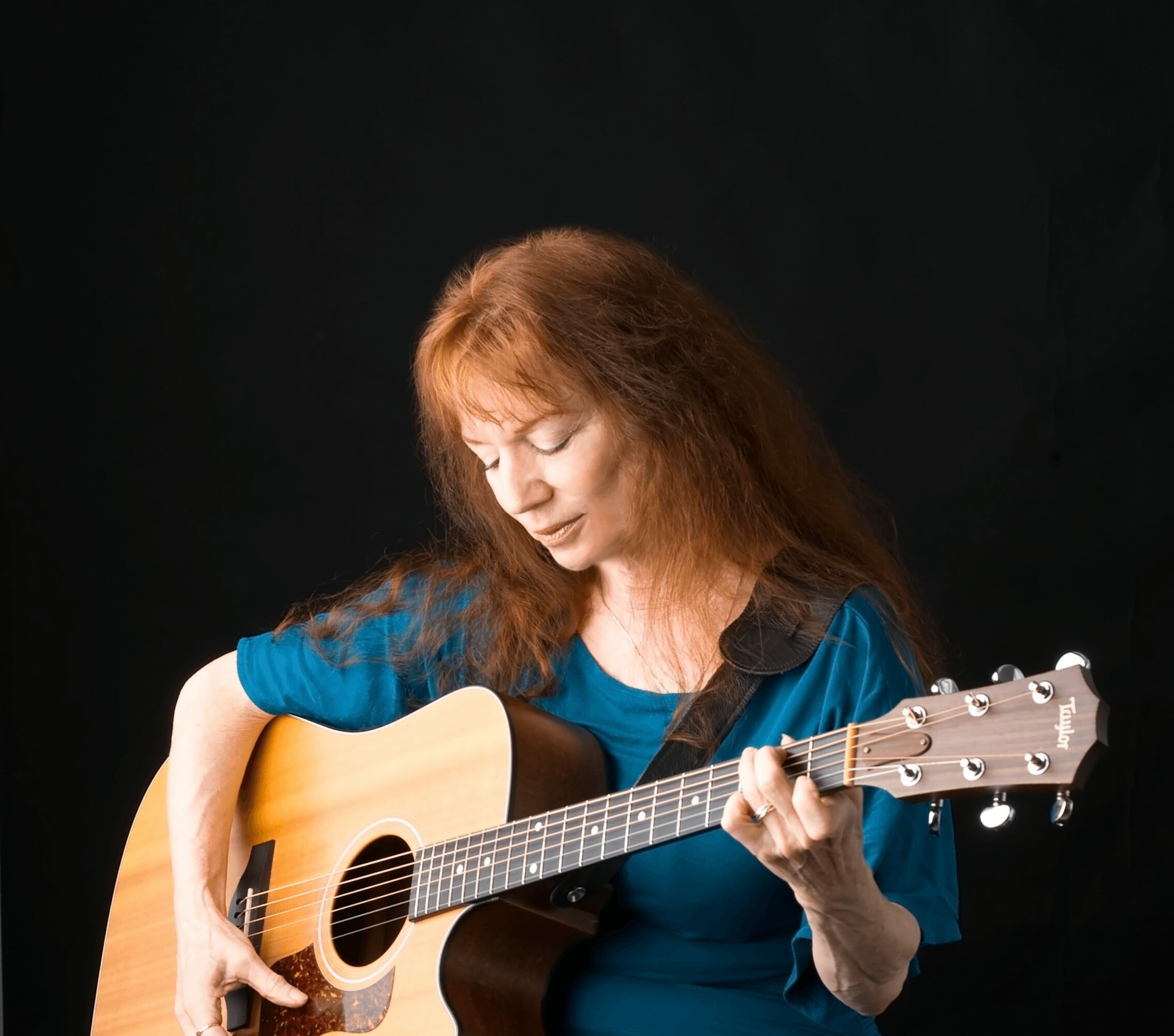 Woman with long red hair playing an acoustic guitar against a black background.