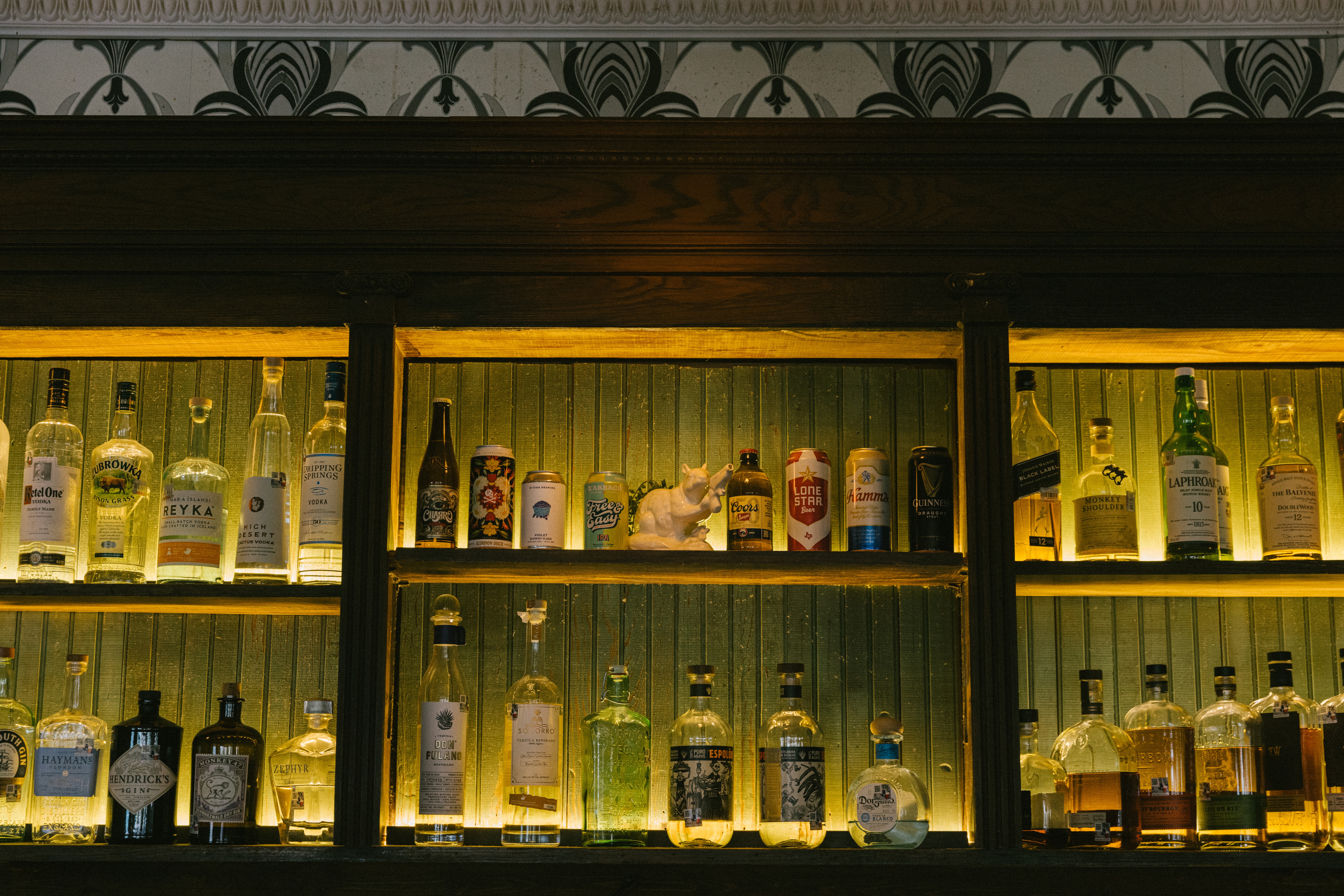 Display of various bottles on shelves with yellow backlighting, arranged in three sections, with a decorative border at the top.