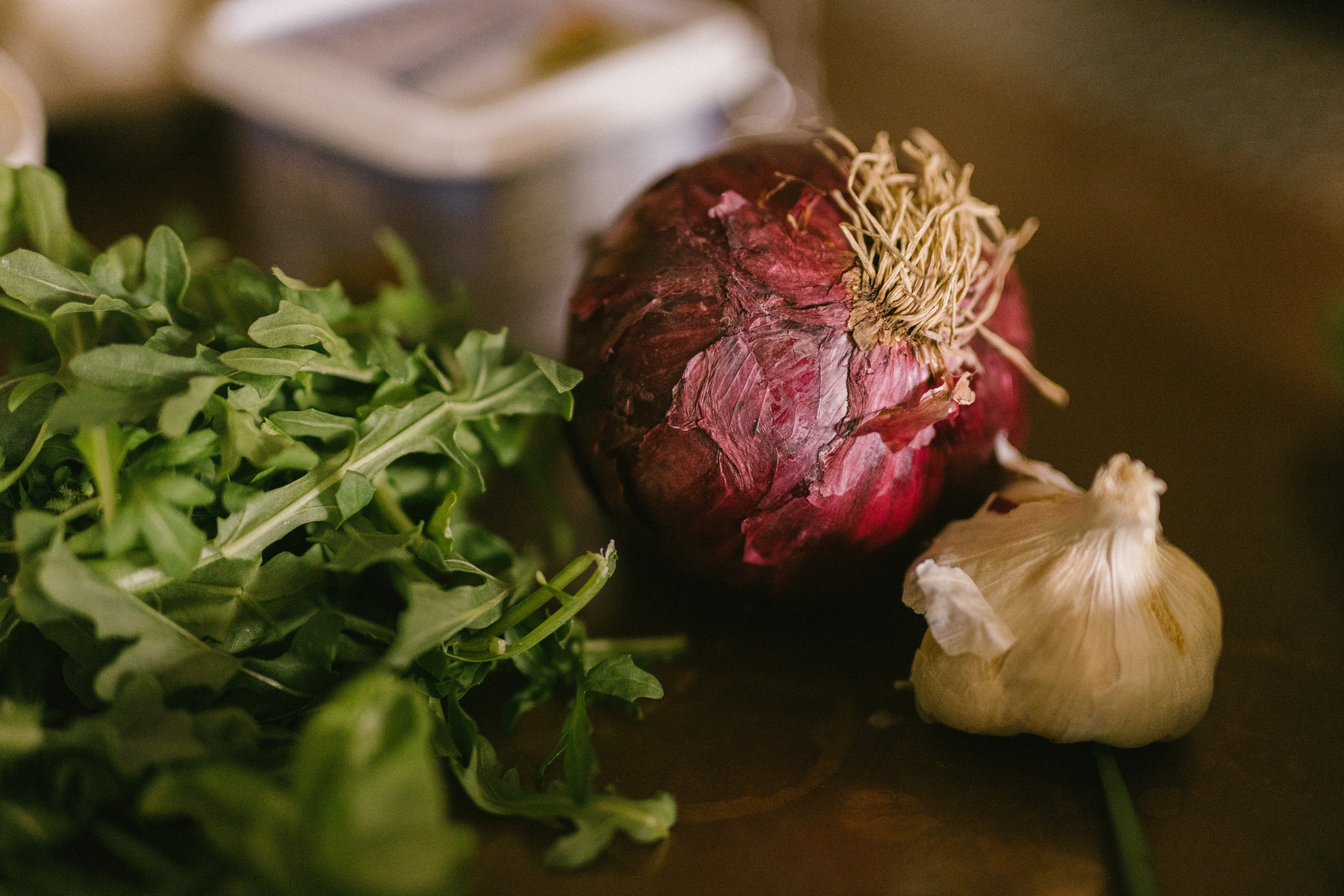 Red onion, garlic bulb, and green leafy herbs on a dark surface.