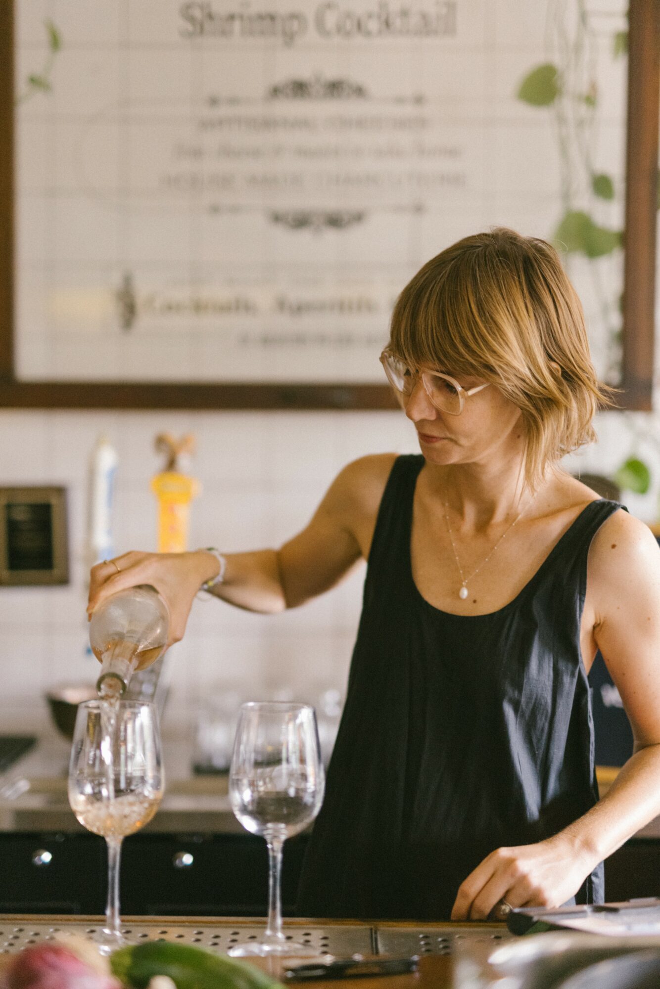 Woman with short blonde hair and glasses pours a drink at a bar, with two glasses in front, blurred menu in background.