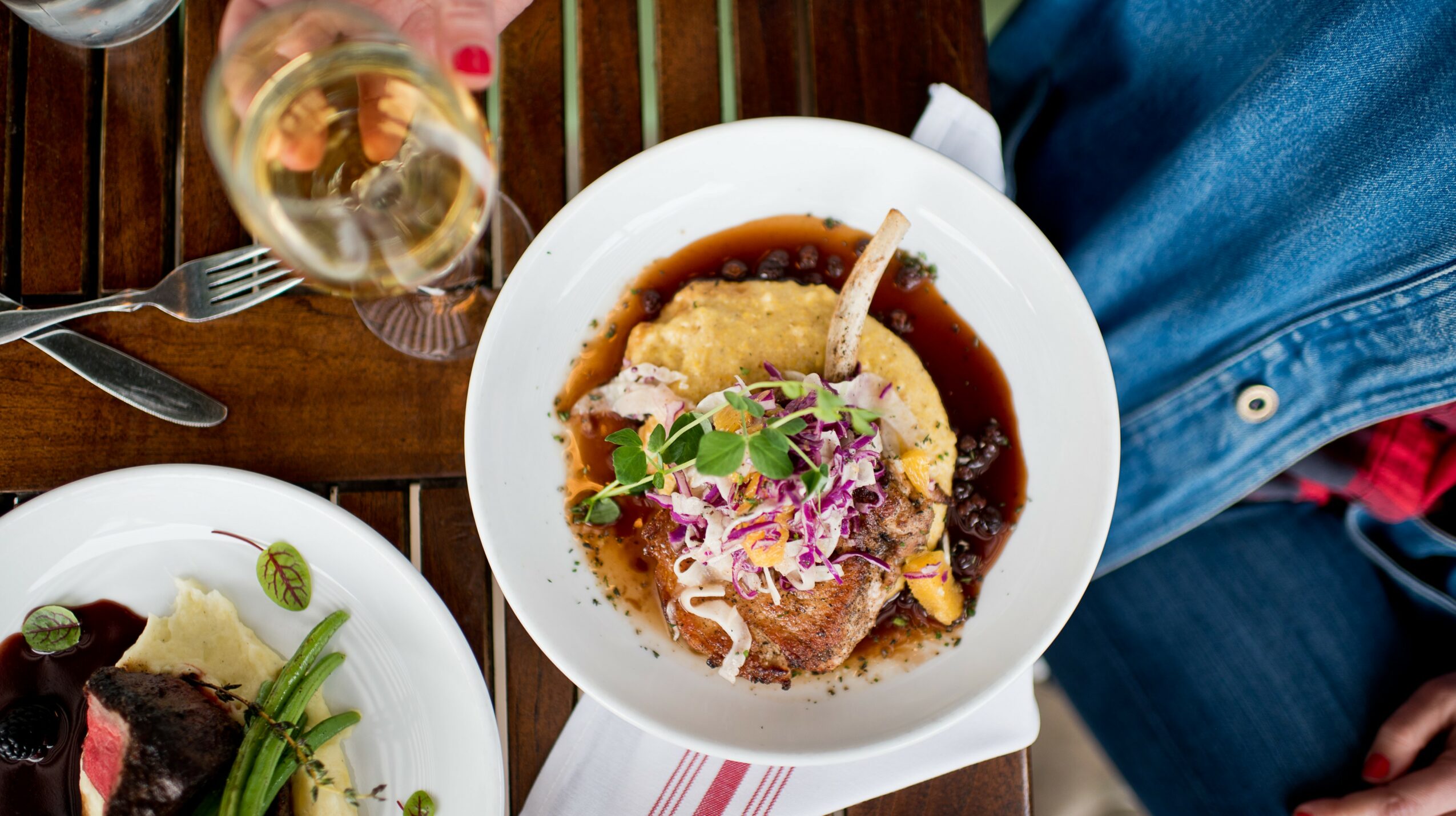 Top-down view of a bowl of food with meat, vegetables, and sauce, on a wooden table with a glass of white wine nearby