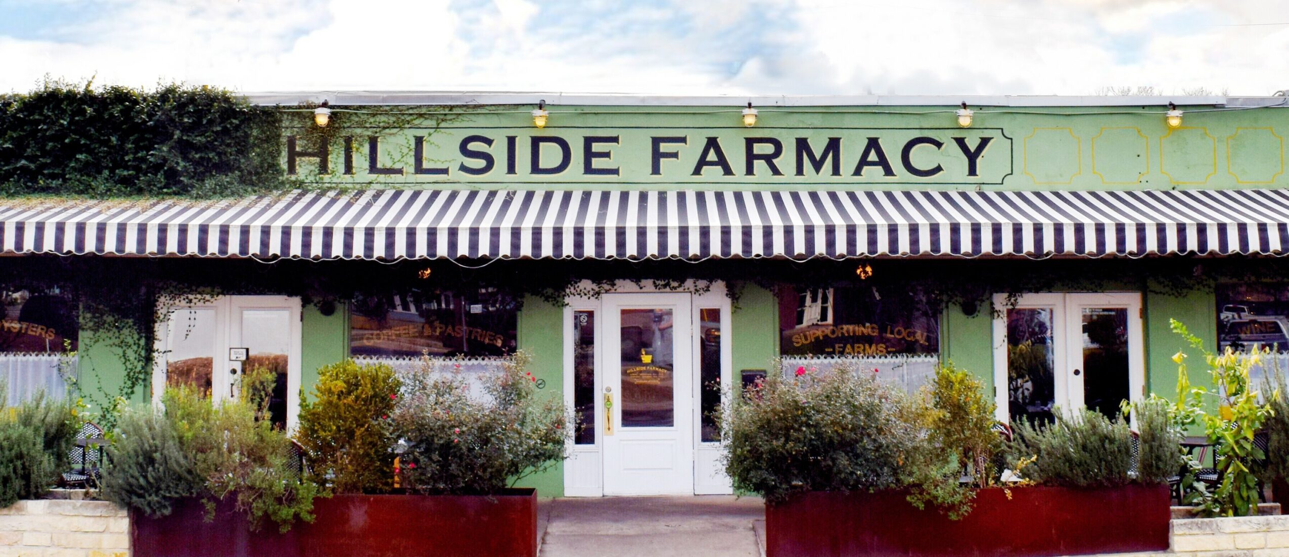 Front view of a green building with a sign reading 'Hillside Farmacy' and plants outside.
