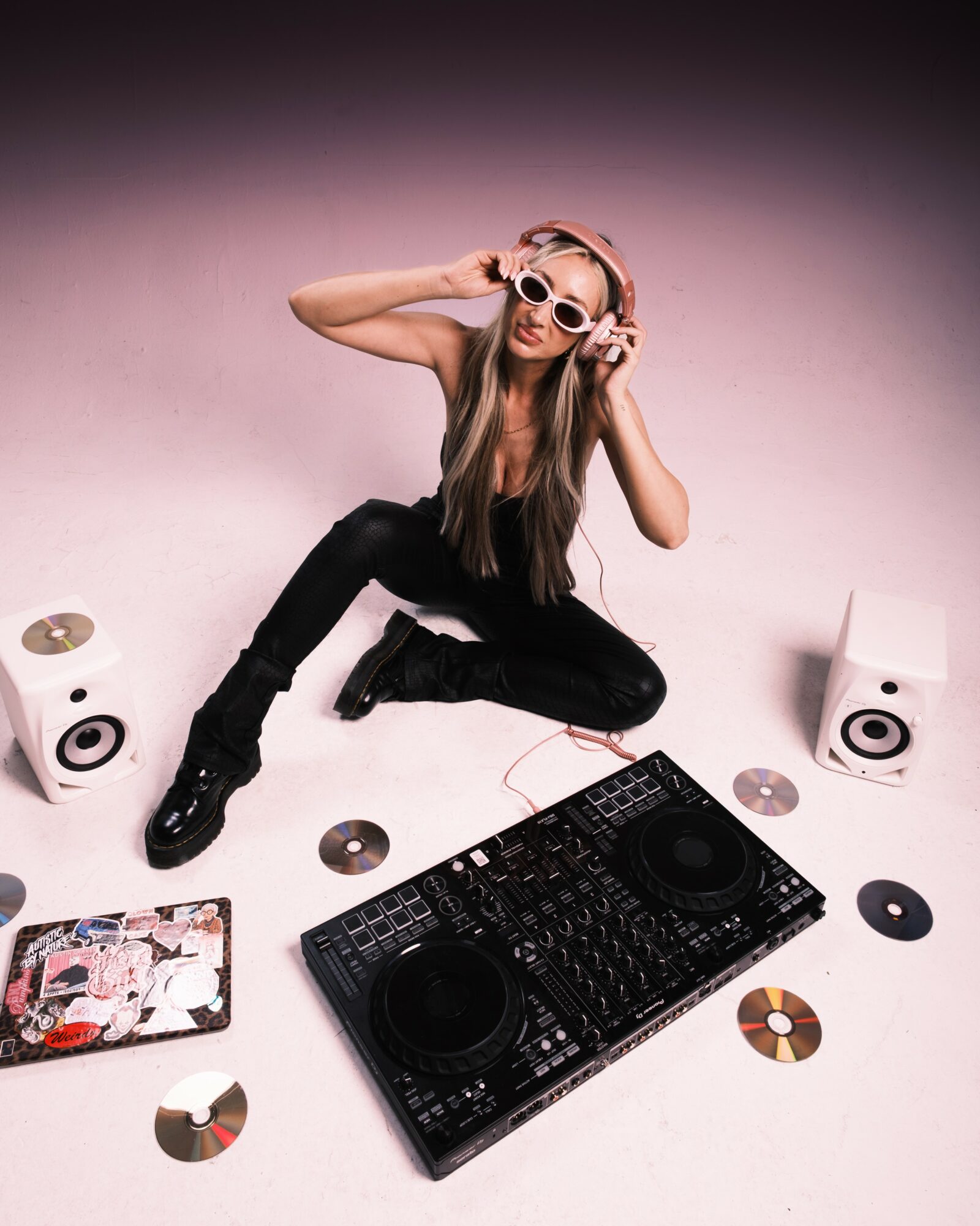 Young woman with long hair, wearing sunglasses and headphones, sitting on the floor with DJ equipment and CDs around her.