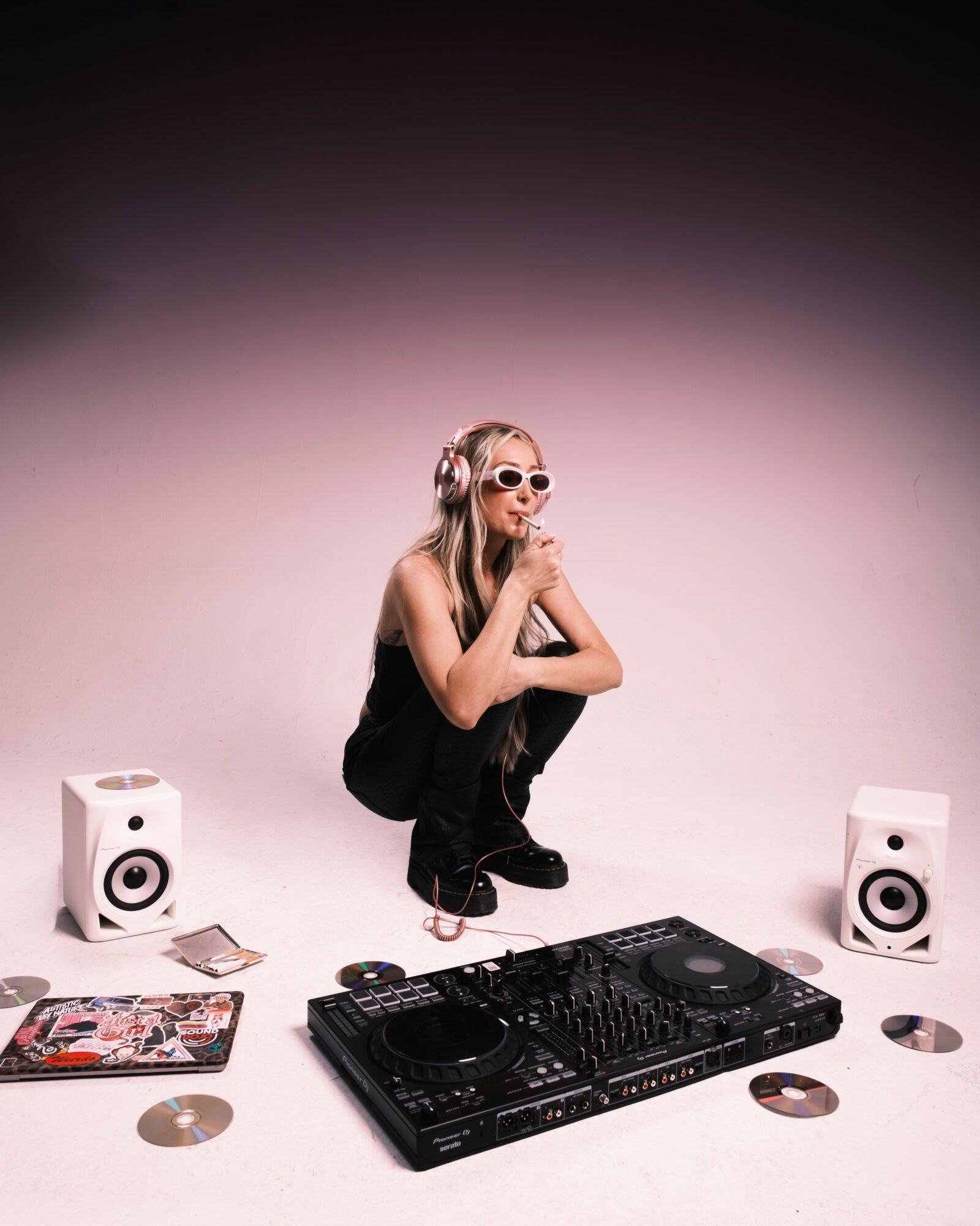 Person crouching with sunglasses, surrounded by DJ equipment and speakers, against a pink gradient background.